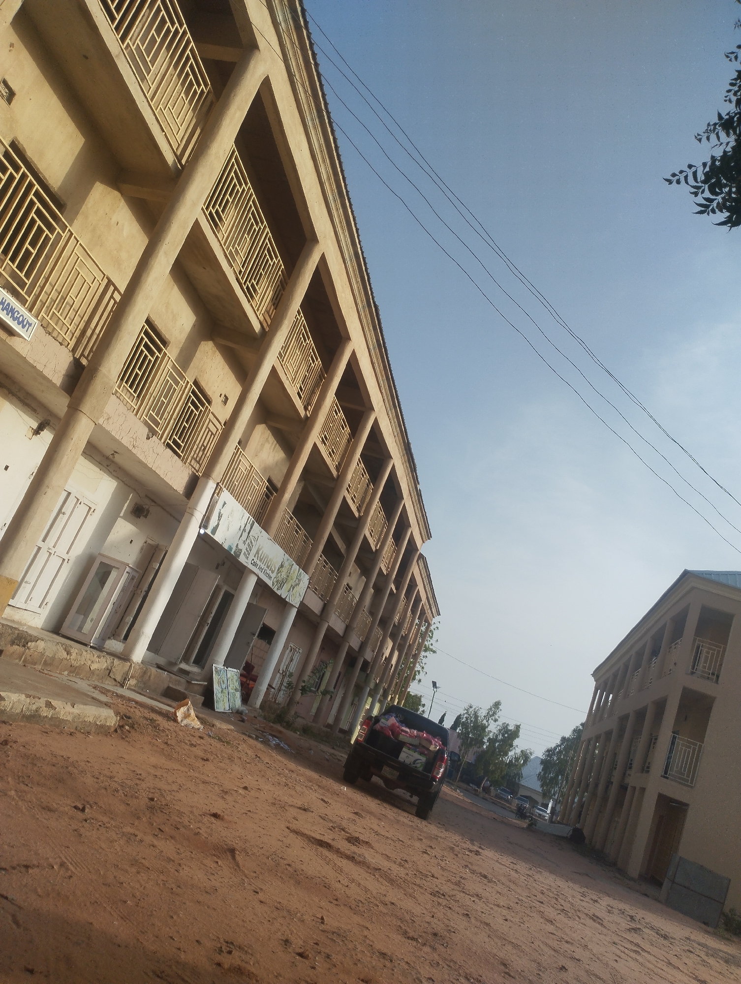 The image shows a street scene in Gombe, Nigeria. Two multi-story buildings flank a dirt road. One building houses a business called "Kumis Cafe And Kitchen." A black pickup truck with items in its bed is parked on the dirt road. Overhead power lines are visible against a clear sky. The scene is slightly tilted, affecting the perspective. Other vehicles are visible further down the street.