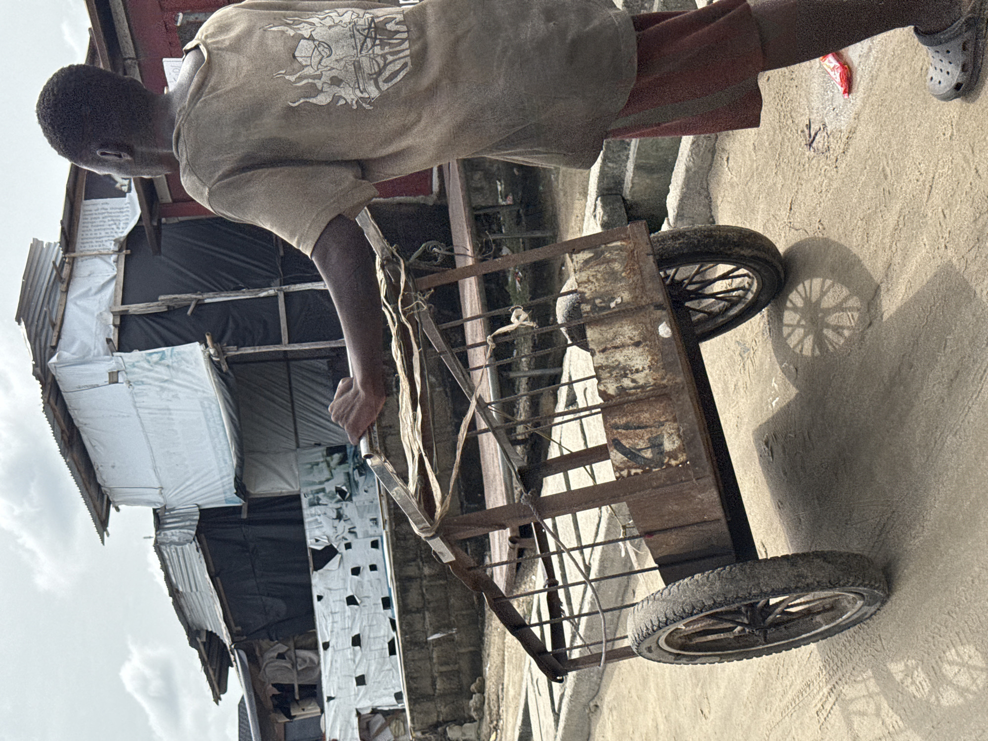 A medium shot, rotated counterclockwise, captures a young person pulling a rusted metal cart along a concrete street. The person is positioned to the left of the cart, their brown-skinned arm extending to grip the handle. They wear a tan t-shirt and shorts, with gray Crocs on their feet. To the left, a structure is visible with walls covered in white plastic and black fabric. The sky is overcast, and the location is in Ado, Nigeria.