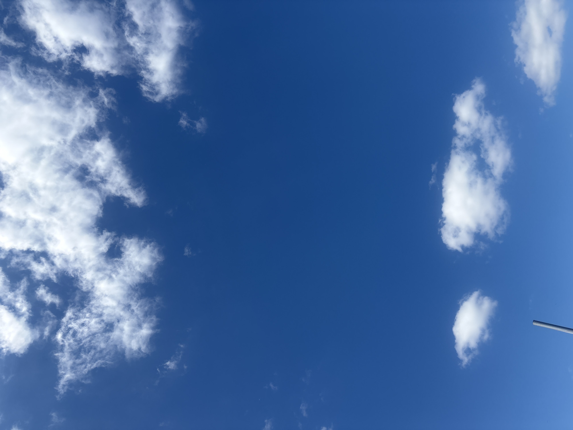 A clear, bright blue sky is the dominant feature of this image, with scattered white cumulus clouds. The clouds are present in various sizes and formations, with larger, more defined clouds towards the top of the frame and smaller, wispy clouds closer to the horizon. In the lower portion of the image, a thin, grey, vertical pole extends upwards from the bottom edge, partially obscuring a few of the lower clouds. There are no people or discernable structures visible, apart from the pole which could be part of a lamppost or flag pole. The scene suggests an outdoor environment on a sunny day, likely in the city of Orsha, Belarus, given the context.