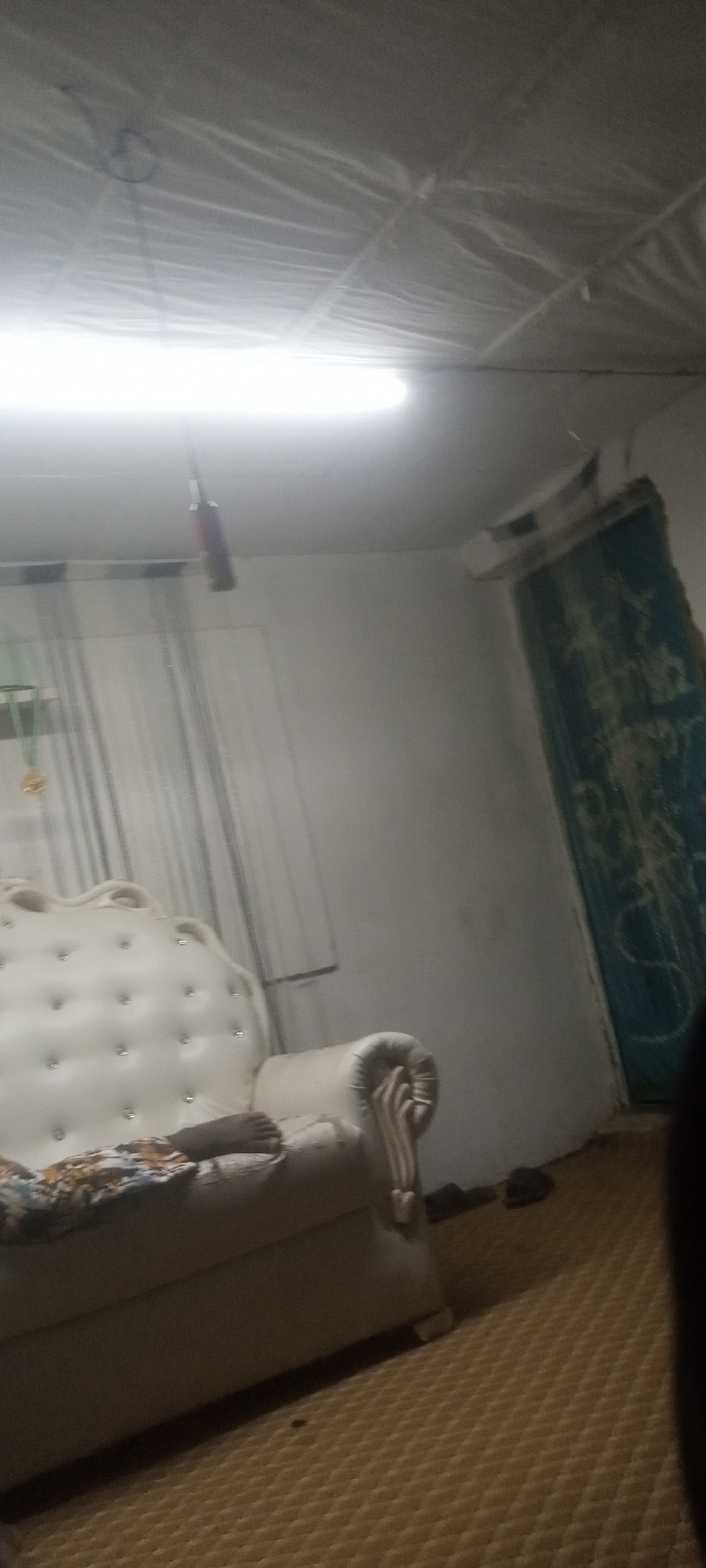 A dimly lit interior scene features a white, tufted sofa with floral patterned fabric. A foot is resting on the sofa cushion. In the background, sheer curtains hang in front of a window. To the right, there is a dark green door or tapestry with intricate patterns, an air conditioning unit above. The ceiling is covered with a white plastic sheeting. A long, bright light fixture illuminates the room. A red, cylindrical object is suspended from the ceiling. The floor is covered with a brown, textured rug. The location is Unknown, Unknown.