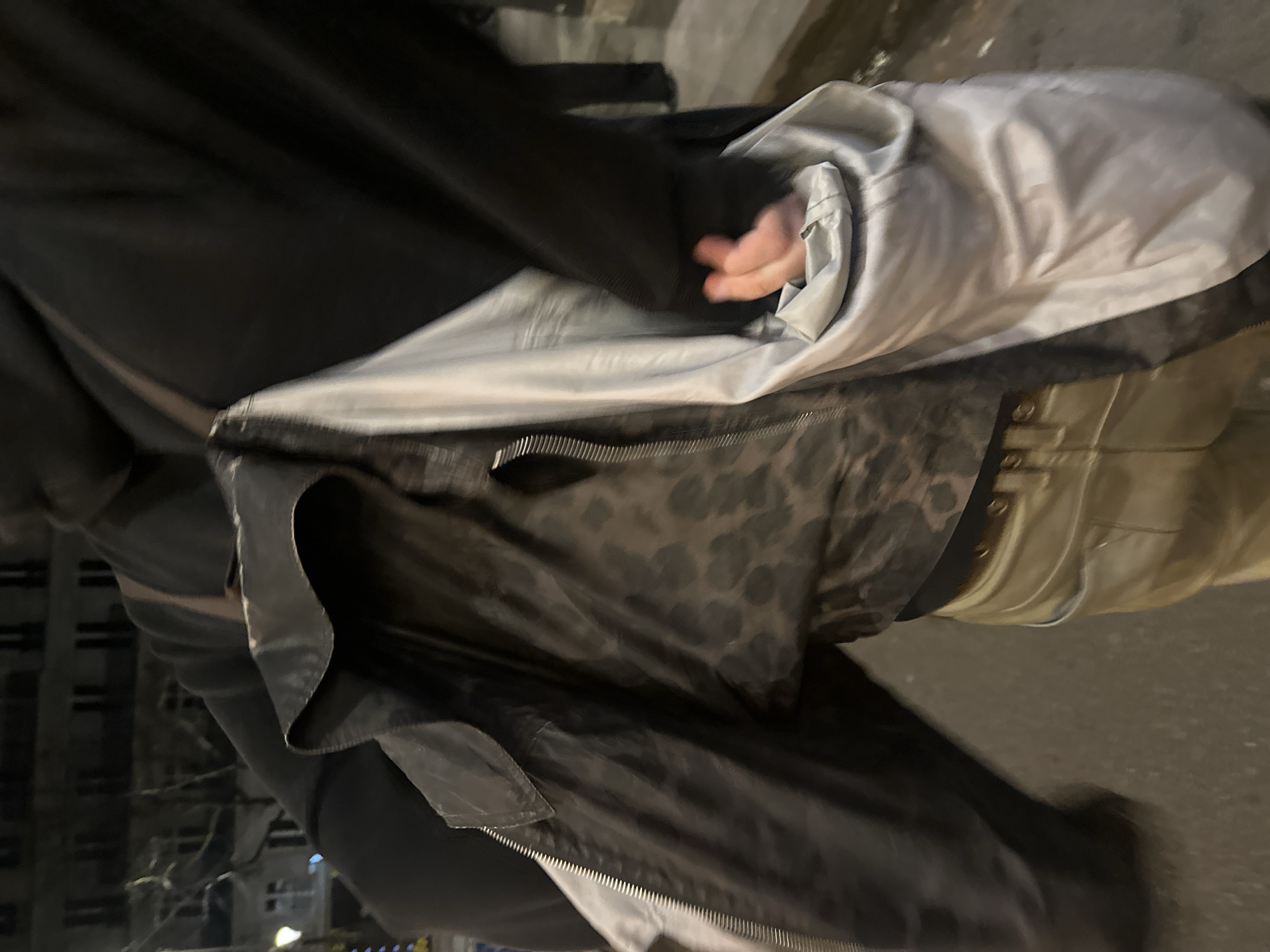 The image depicts a nighttime urban scene in Paris, France, focusing on the mid-torso of an individual from a slightly low perspective. The person is wearing a dark, unzipped jacket, revealing a light grey or silver inner lining. Beneath the open jacket, a dark garment with a subtle, mottled, or leopard-print pattern is visible, worn over a black hooded garment. A brown strap, indicative of a bag or backpack, passes over the individual's left shoulder. The person's right hand is partially curled and positioned within the light-colored inner lining of the jacket. The lower visible clothing consists of light brown or khaki pants, secured by a brown belt with a visible buckle. The background features dark, multi-story buildings with characteristic Parisian architecture, including distinct window patterns, silhouetted against the night sky. A streetlamp with a yellowish glow is visible on the far left, and bare tree branches are discernible against the darkness. The ground surface appears to be paved.