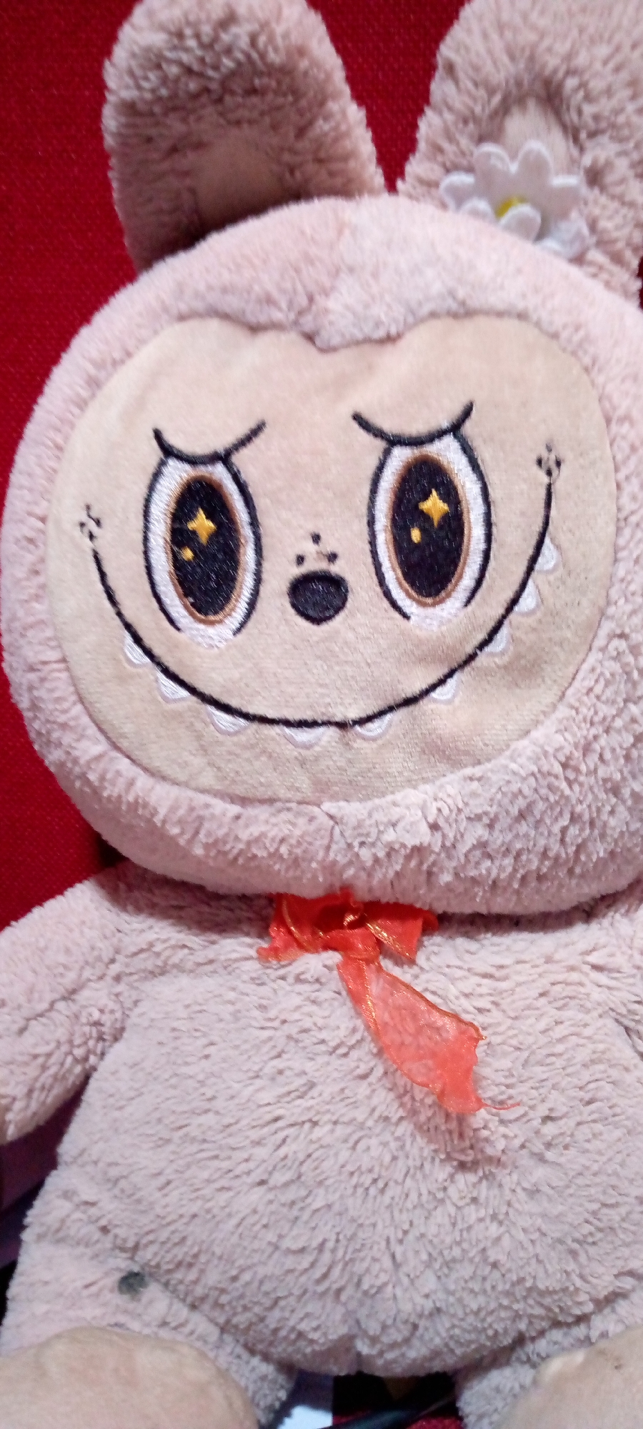 A close-up shot captures the face of a pink plush toy, likely a stuffed animal, against a red background. The toy has large, embroidered eyes with black pupils and golden star-shaped highlights, giving it a whimsical appearance. Its mouth is depicted as a wide, stitched smile with a row of small, white, embroidered teeth. A tiny black nose sits above the smile, and two small embroidered dots on each cheek complete its facial features. On its head, two large, soft ears protrude upwards, and a small white flower with a yellow center is attached to the right ear. A bright orange ribbon is tied in a bow just below the toy's chin. The plush material of the toy is soft and fuzzy, with a light pinkish-beige hue. The background is a solid, textured red, suggesting it might be a fabric or a painted surface. The lighting in the image is even, highlighting the details of the toy's face and texture.