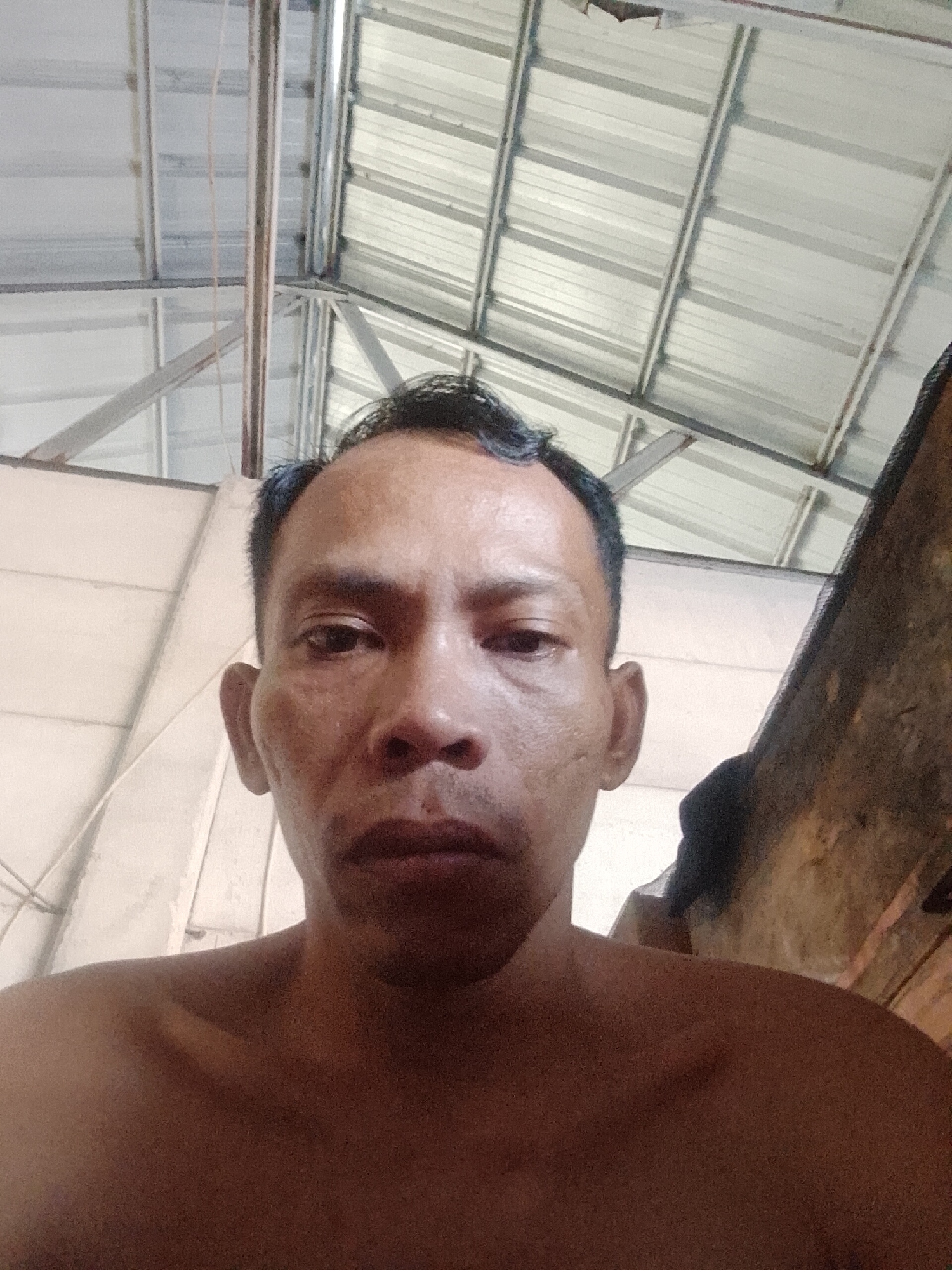 A close-up, slightly angled, eye-level shot features a man's face and upper torso against a cluttered background. The man has dark hair, a weathered complexion, and is shirtless. The background shows a corrugated metal roof structure supported by a metal framework. To the right, an unidentifiable object is visible. The image was taken in Bukit Ketok, Indonesia.