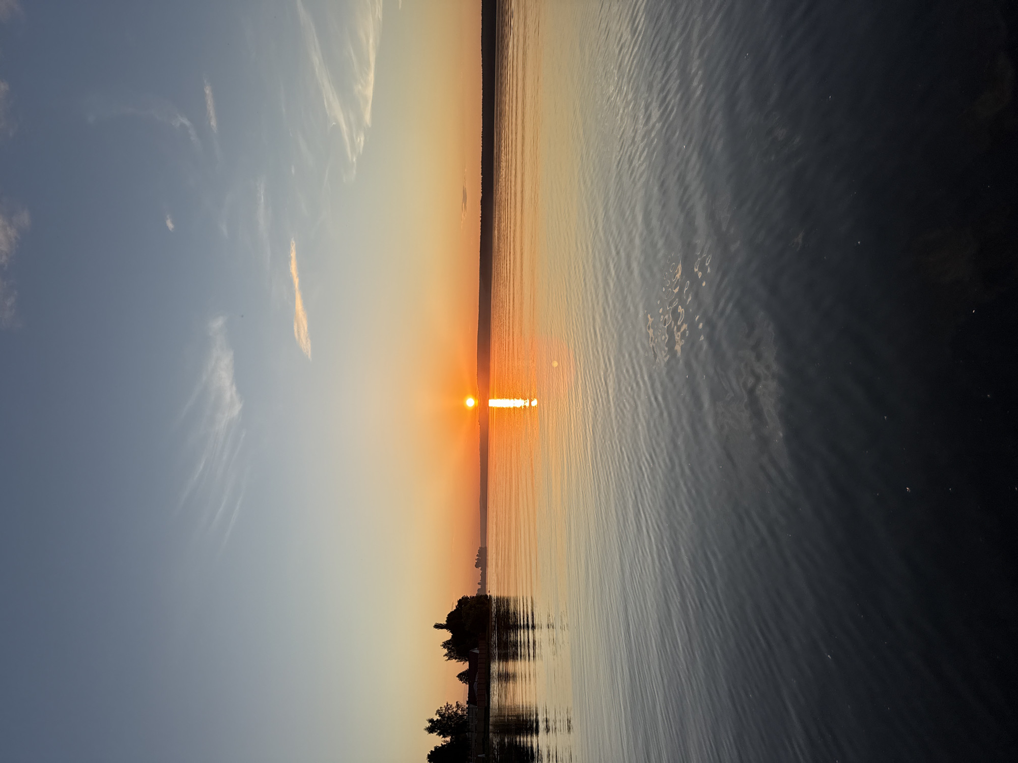 The image displays a serene sunset over a vast body of water, likely the Votkinsk Reservoir, in Votkinsk, Russia. The sun, a distinct bright orange orb, is positioned low on the horizon, slightly to the right of the central axis. A prominent, elongated vertical reflection of intense orange and yellow light stretches directly below the sun across the water's surface. The sky transitions from a pale blue at the zenith to warm orange and yellow tones closer to the horizon, with sparse, wispy cirrus clouds distributed throughout. The water's surface is characterized by gentle, consistent ripples, reflecting the sky's gradients and appearing dark blue-grey in the foreground, lightening to warm hues towards the horizon. The distant left shoreline features a dark silhouette of various trees and several structures, including a building with a red roof and a more prominent dark structure, possibly a church or large house, behind a visible fence line. The distant right shoreline presents a low, dark band of land, likely forested, extending to the horizon. No individuals are visible in the scene.