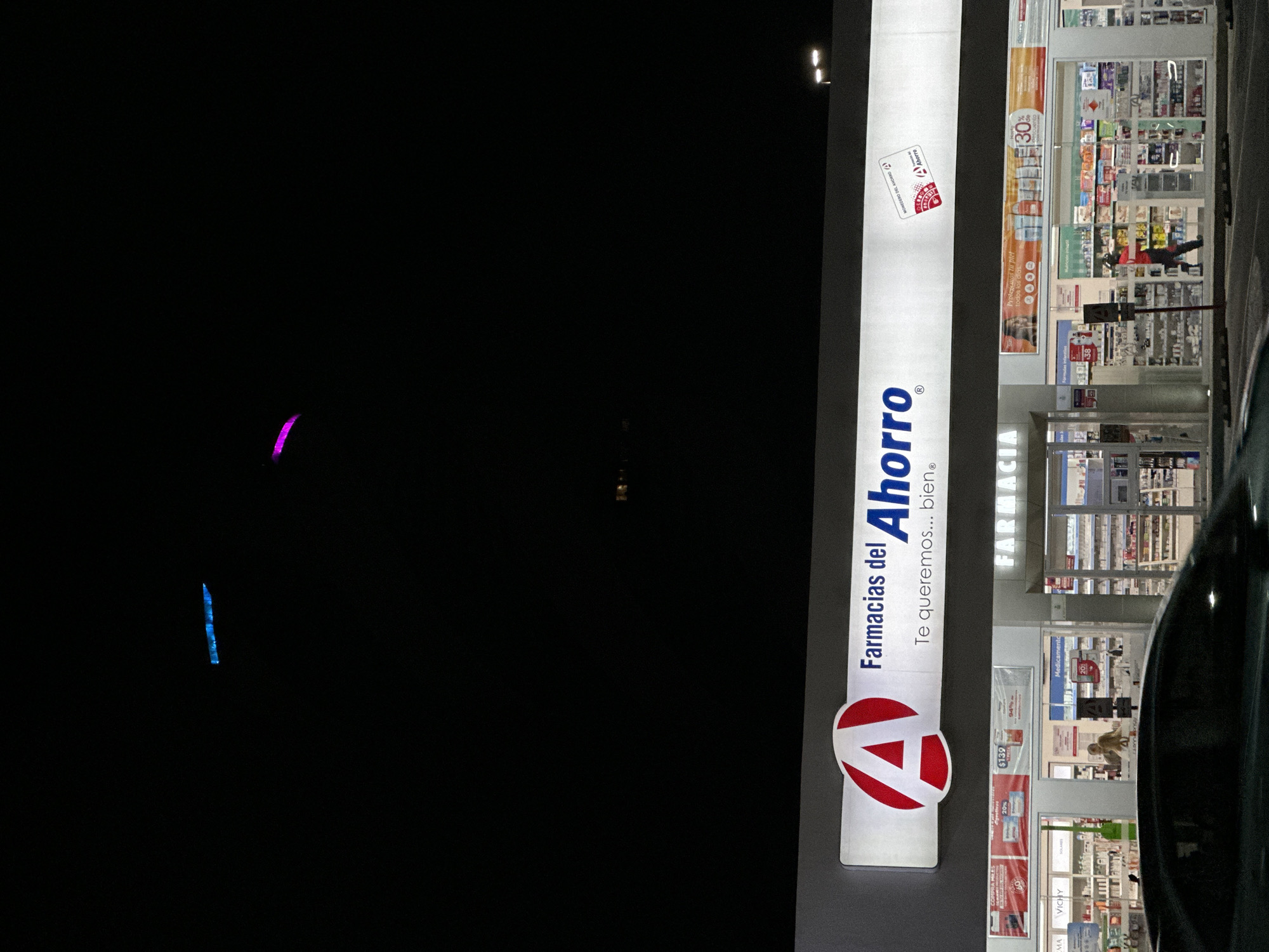Here is a description of the image:
The image depicts a "Farmacias del Ahorro" (Savings Pharmacy) at night. The pharmacy's illuminated sign, with the company logo and the tagline "Te queremos... bien" (We love you... well), is prominently displayed. The pharmacy's interior is visible through the windows, showing shelves stocked with products and promotional materials. There are hints of light in the dark night sky, with blue and purple neon lights. Parts of a dark car are visible in the reflection.