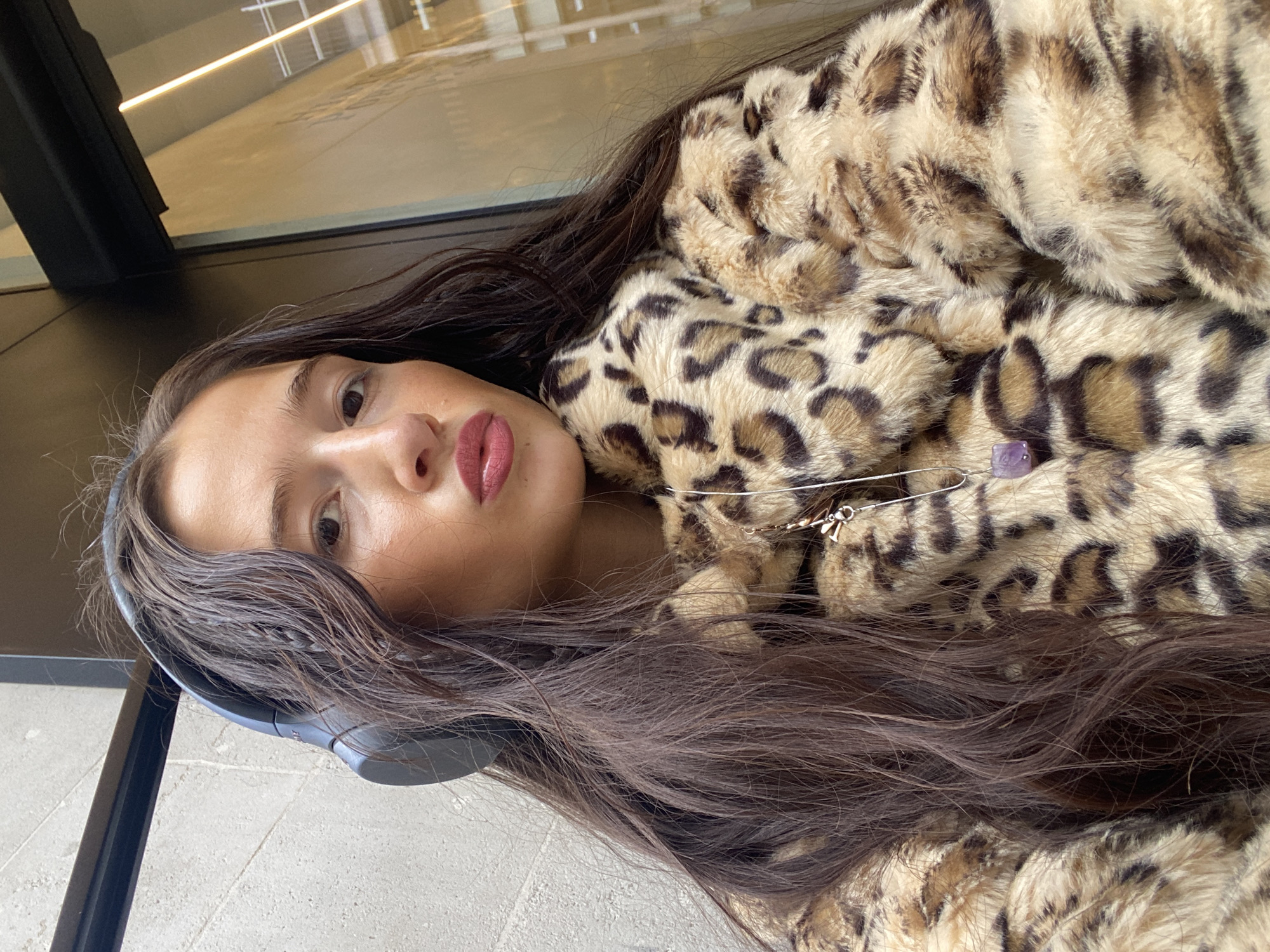 A close-up portrait shows a young woman looking directly at the viewer, with her head resting sideways or tilted. Her long, dark brown hair cascades around her face and across a faux fur coat. She has full lips, enhanced with a reddish-pink lipstick, and her expression is neutral. Dark grey over-ear headphones are visible on her left ear.
She is wearing a plush, light brown faux fur coat with dark brown leopard print patterns. Pinned to the coat is a silver accessory, possibly part of a necklace or a decorative pin, featuring a small metallic charm and a small, translucent purple crystal bead, which appears to be amethyst.
The background is subtly blurred, revealing a light grey, possibly concrete or tiled, floor in the lower part of the image. A dark, angular structure, possibly a window frame or railing, runs along the left side. In the upper background, there's a reflection on a large glass surface, showing linear light fixtures or architectural elements, suggesting an indoor, modern setting such as an airport, office building, or shopping center.
The woman appears to be posing for a photograph or selfie. The time of day seems to be daytime, indicated by the natural light implied by the reflections. There is no visible text in the image. The provided location context is London, United Kingdom.