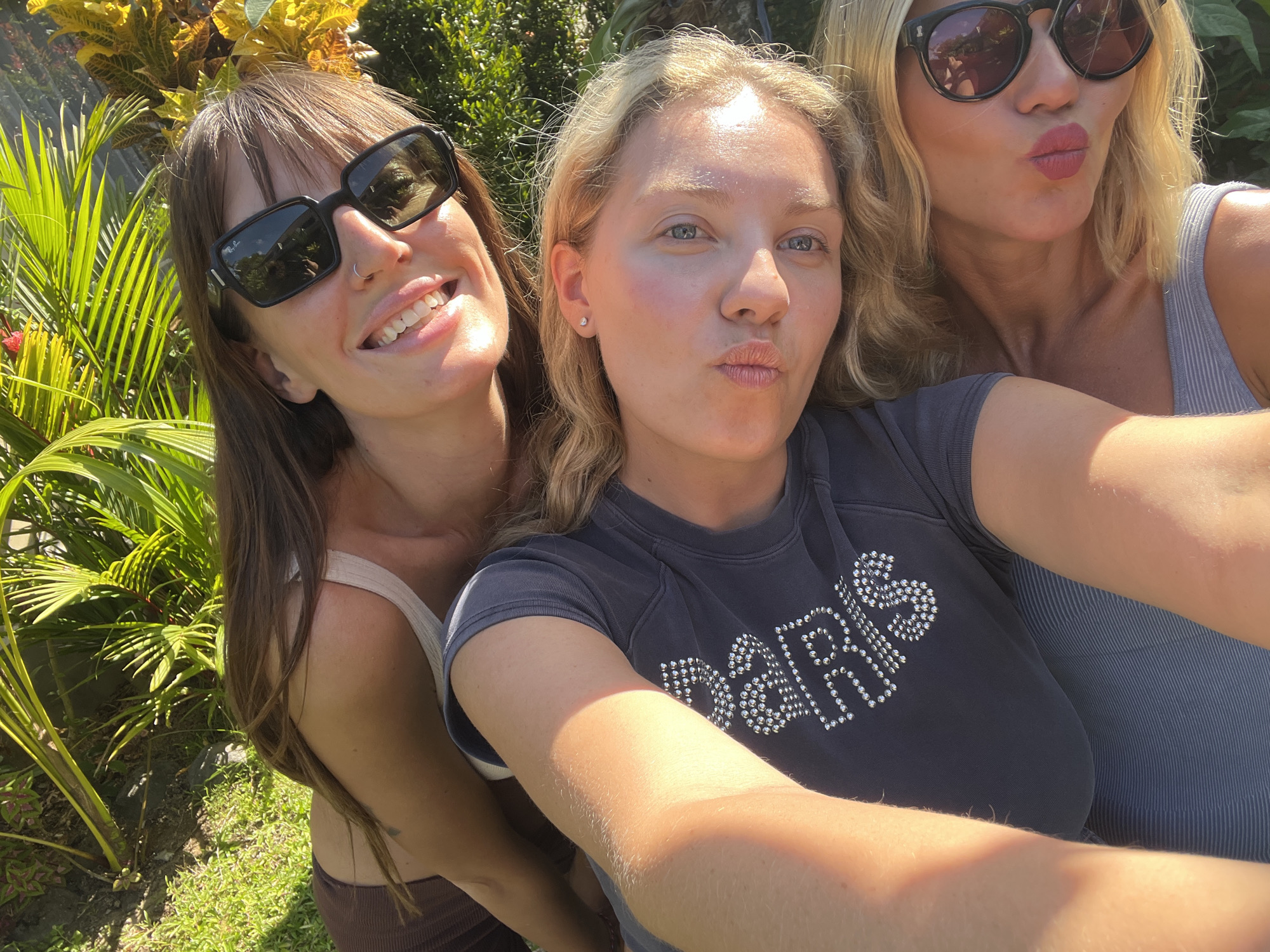 The image shows three light-skinned women posing for a selfie outdoors in North Kuta, Indonesia. The woman on the left wears sunglasses and has brown hair. The woman in the center has blonde hair and is wearing a dark gray shirt. The woman on the right also has blonde hair, sunglasses, and is wearing a gray tank top. All three women are pursing their lips. Tropical foliage is visible in the background. The ground is covered in green grass.