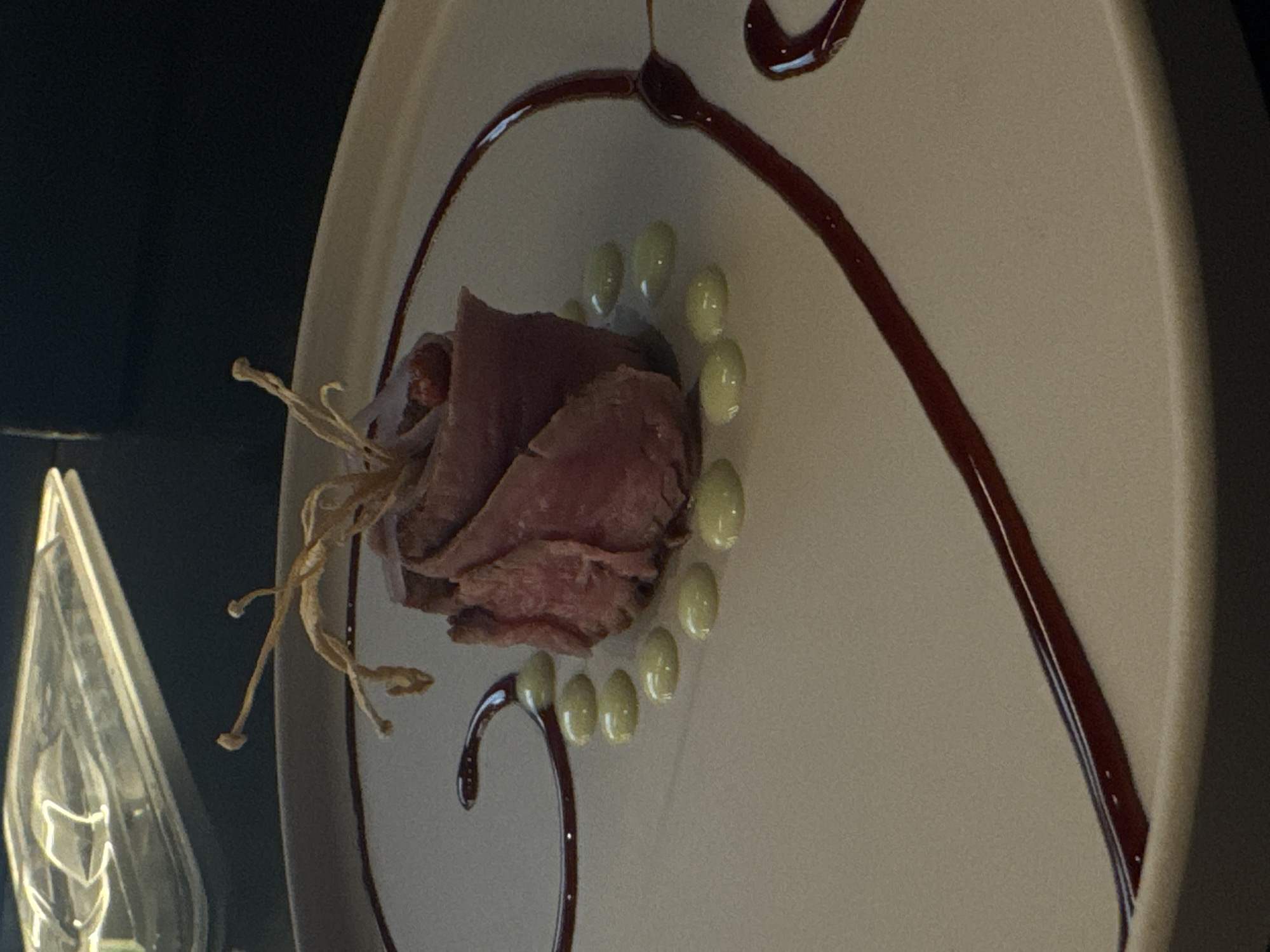 This image shows a close-up of a beautifully plated dish on a light-colored ceramic plate. The main subject is a piece of thinly sliced meat, expertly rolled and arranged to resemble a rose. It is garnished with dried, delicate tendrils at the top, one of which has a small, red berry or bead. Surrounding the meat rose are several small, pale green dollops of sauce, arranged in a semi-circular pattern. A dark, rich red sauce is artistically swirled across the plate, creating elegant lines and curves. The background is dark and out of focus, with a hint of a reflective, metallic-looking object in the upper left corner, possibly a lid or a decorative element. The lighting suggests an indoor setting, likely a restaurant, emphasizing the food presentation. The image does not provide any specific clues or information about the location in the city of Sofia, Bulgaria, or any people present.