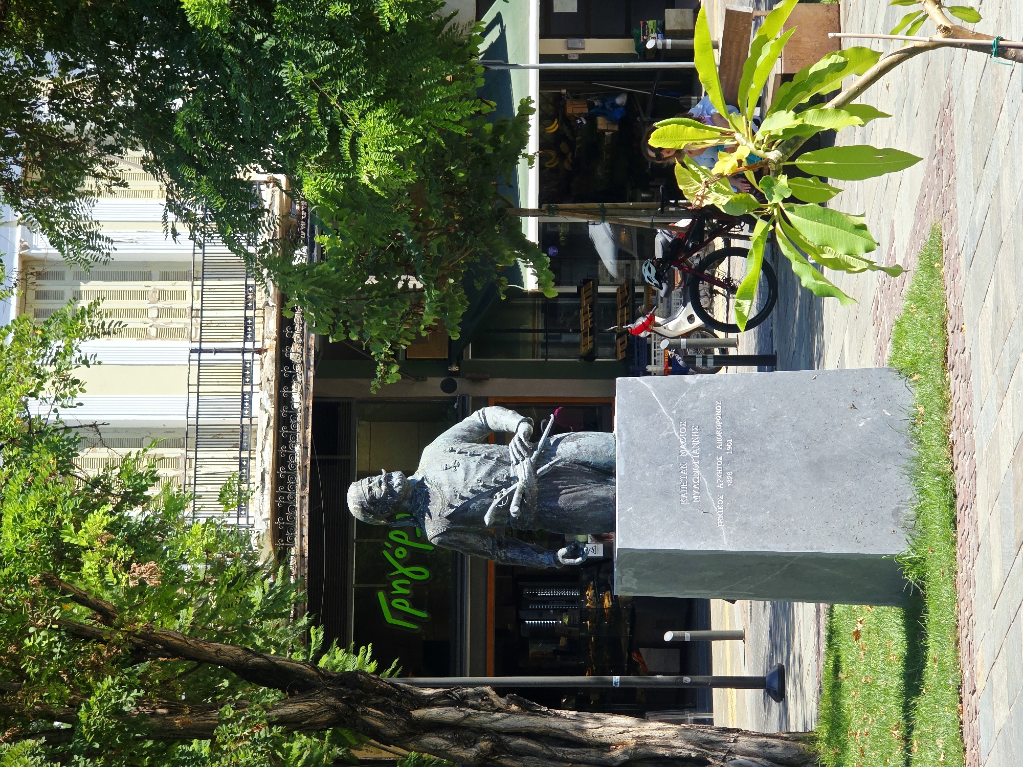 In Chania, Greece, a bronze statue of a man in military attire, possibly historical, stands on a stone pedestal in front of a building. The statue depicts a man with a prominent mustache, wearing a uniform and holding a rifle. The pedestal bears Greek inscriptions. Behind the statue, a store with a green awning and large windows displays various goods, including what appears to be food items. Trees with lush green foliage provide shade, and a section of pavement is visible in the foreground. A woman is seated on a bench to the right, and a motorcycle is parked nearby. The overall scene suggests a public square or street in a Mediterranean city.