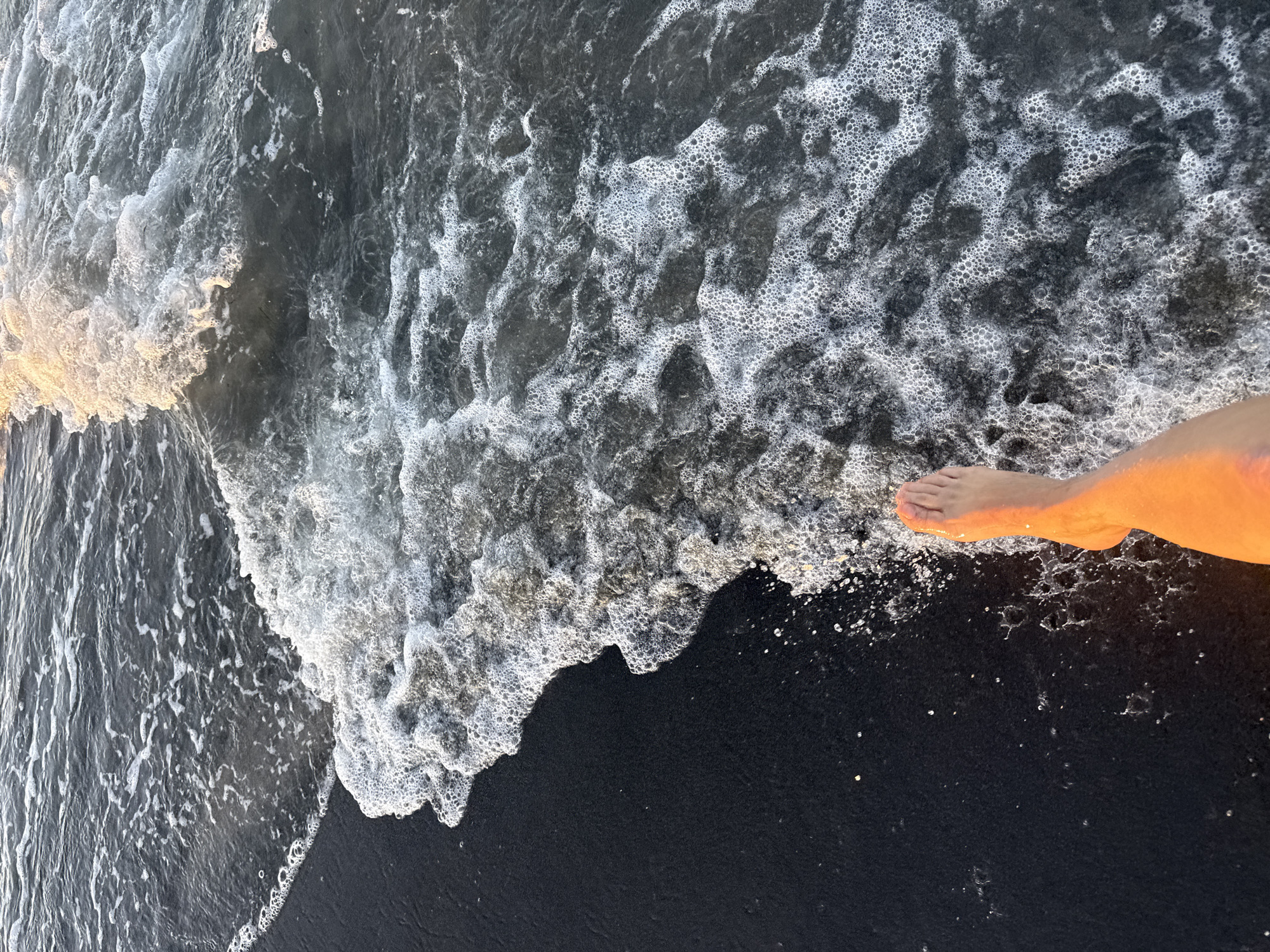 An eye-level, medium shot captures a person's bare foot stepping into the dark sand at the edge of the ocean. The water is dark and foamy, with gentle waves lapping at the shore. The sun, low in the sky, casts an orange glow on the person's leg and foot, creating a warm contrast with the cool tones of the water and sand. This scene is likely taking place on one of Burgas, Bulgaria's many beaches, possibly during the golden hour, a time of day known for its soft, warm light.