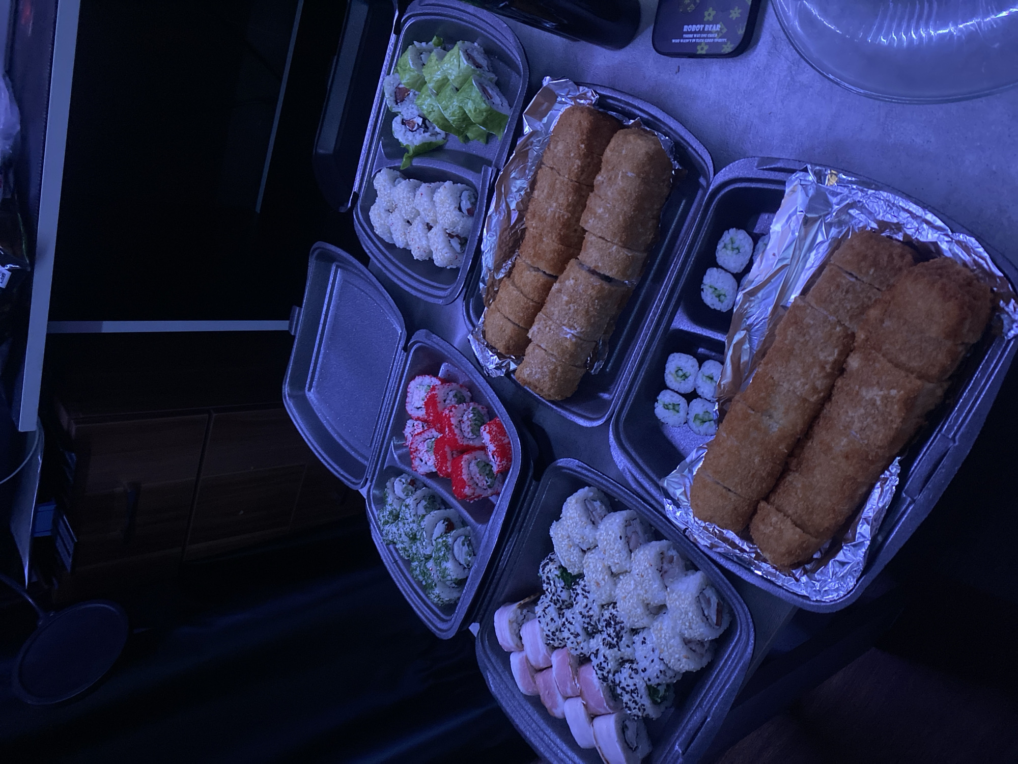 The image shows several take-out containers of sushi arranged on a surface. The containers are made of gray plastic or foil, and they hold various types of sushi rolls. Some rolls are covered in sesame seeds, others have a breaded, fried exterior, and some are topped with colorful roe. In the background, there is a phone case with the words "ROBOT BEAR" visible. The location of the image is unknown and cannot be determined from the visible context, so the claim it is from Izhevsk, Russia cannot be substantiated.