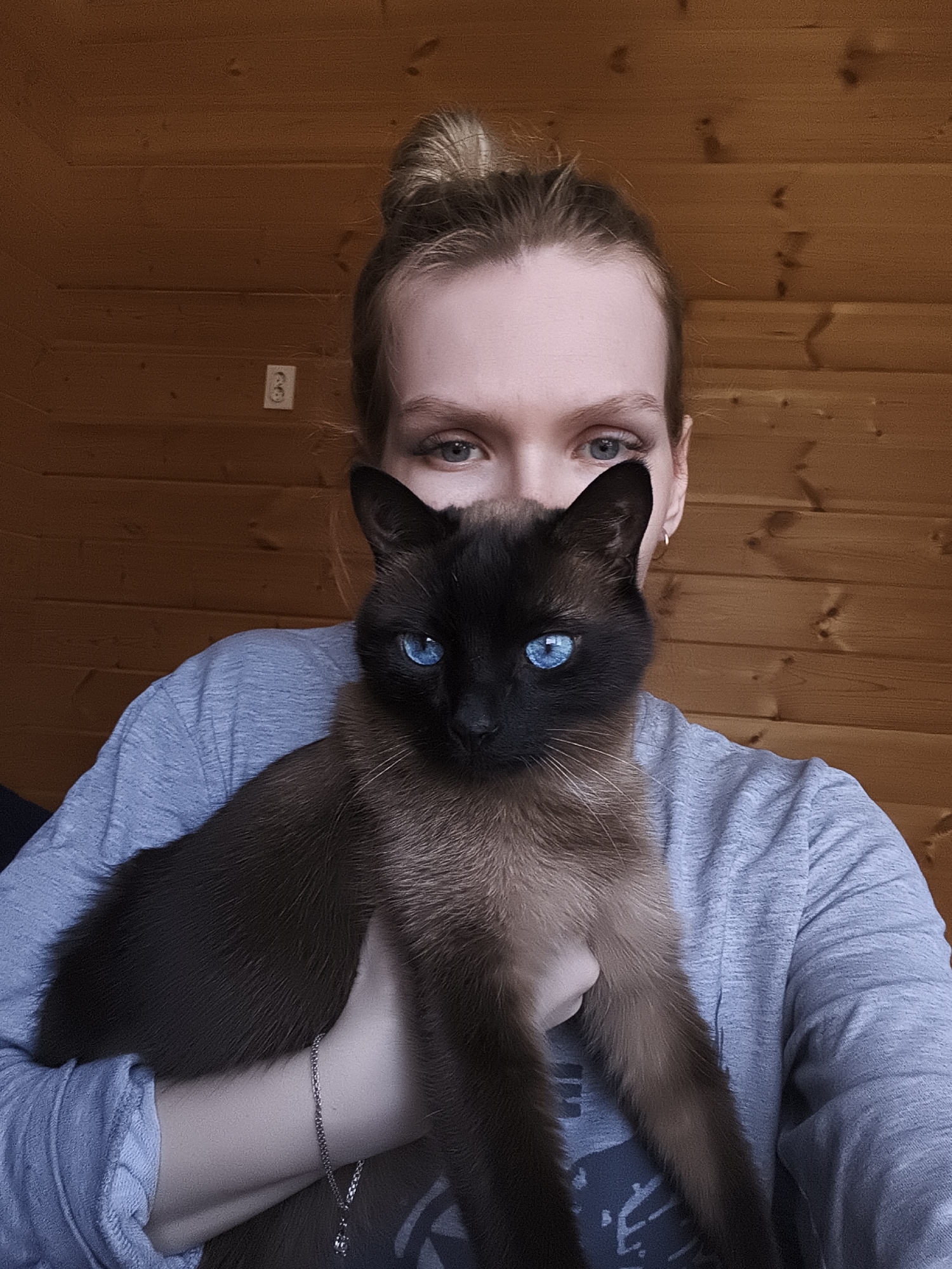 A person with blonde hair pulled back in a bun, blue eyes, and wearing a grey long-sleeved shirt is holding a Siamese cat with striking blue eyes. The background is a wood-paneled wall with an electrical outlet. There is a silver chain necklace around the person's neck. The location is identified as Русский Вожой, Russia.