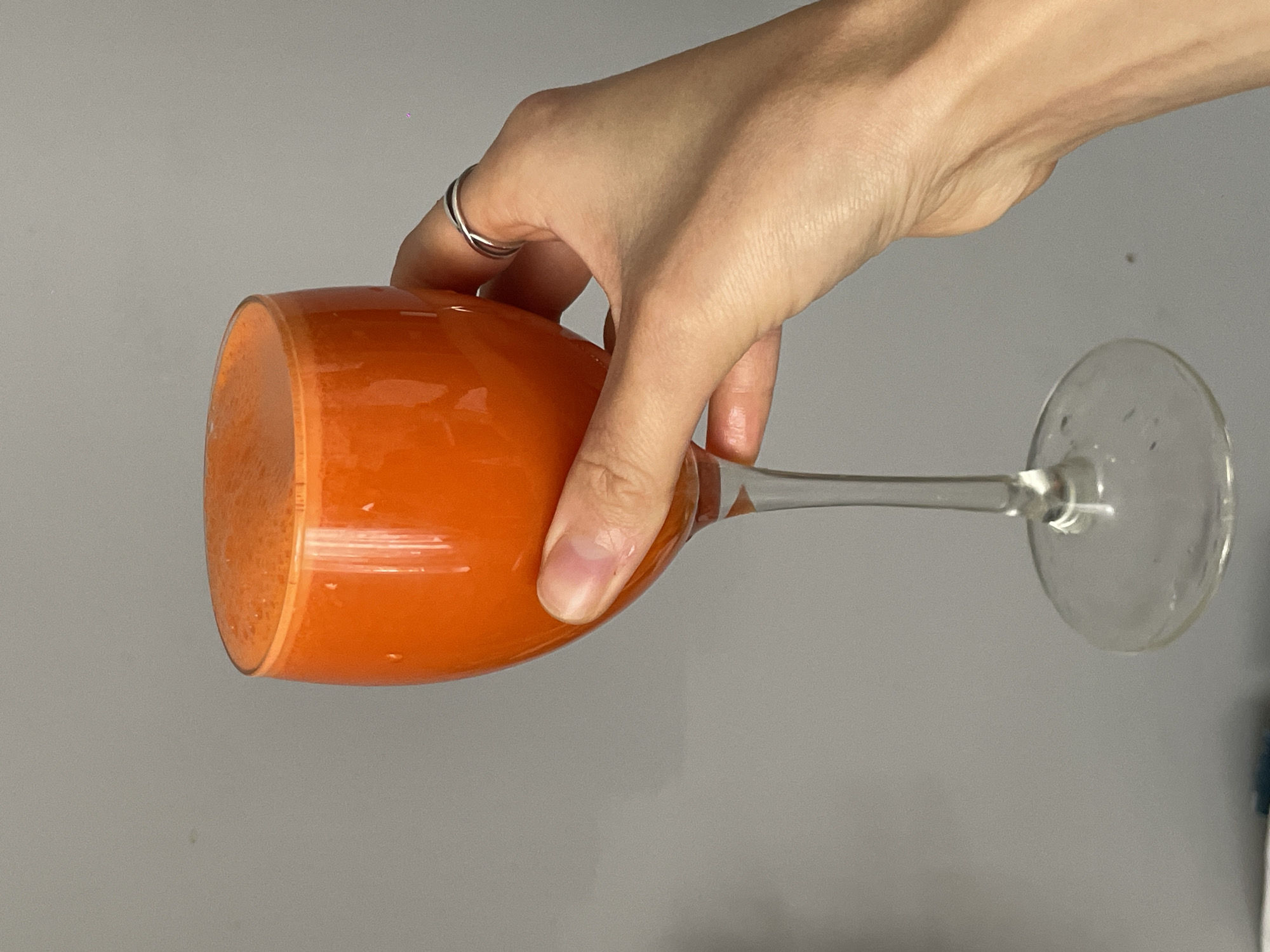 A close-up, horizontal shot shows a person's hand holding a wine glass filled with a bright orange liquid, possibly juice. The hand, with a silver ring on one finger, grips the glass near its base. The wine glass has a clear stem and base. The background is a plain gray wall. The image was taken indoors, and there is no indication of it being in Sochi, Russia.