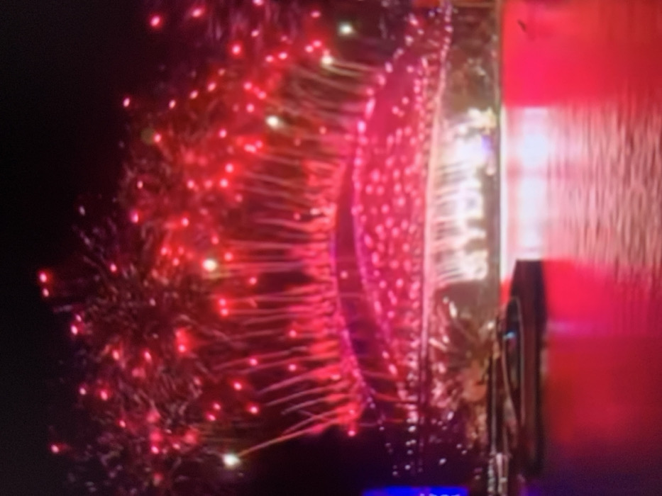 The image shows fireworks in the night sky, with many red sparks and streaks emanating from a central point. Part of a building or structure is visible on the right, with a red panel and reflective surface. A small display screen with a blue glow is at the bottom. The overall scene is dark, with the bright fireworks providing the main source of illumination.