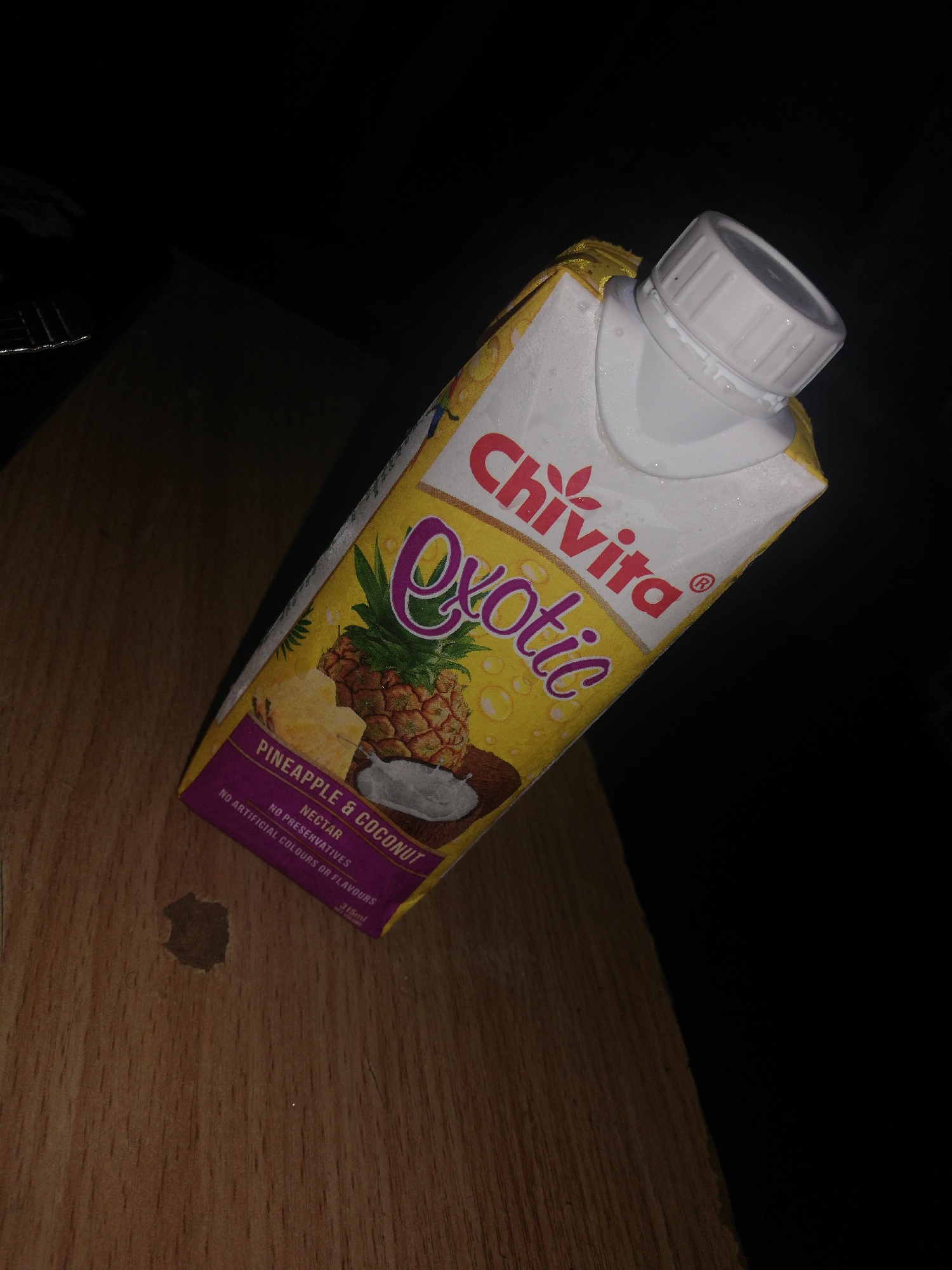 The image shows a 315ml carton of Chivita Exotic pineapple and coconut nectar on a wooden surface. The carton is upright, with a white plastic cap visible. The packaging is yellow with an image of a pineapple and coconut. The lower part of the carton is purple with the product description "Pineapple & Coconut Nectar" and "No artificial colours or flavours". There's a small dark stain on the wooden surface near the carton. The environment is dim, with a dark background. Location information is unavailable.