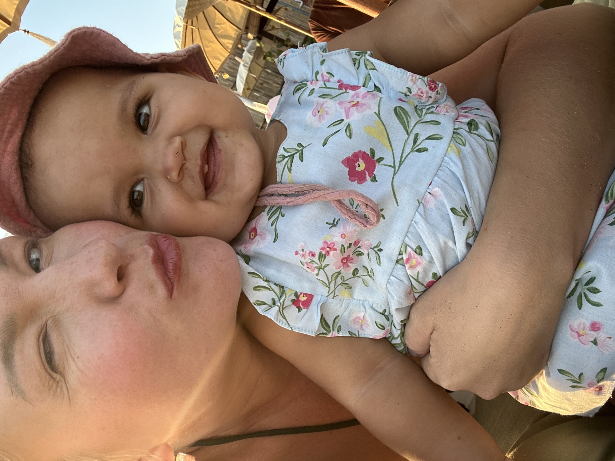 A close-up, low-angle shot shows a woman on the left, winking with her lips puckered, as she embraces a baby. The woman has blonde hair, fair skin, and is wearing a dark green swimsuit strap. She appears to be at a beach, evidenced by the sandy texture on the baby's arm and dress, and the presence of beach umbrellas in the background. The baby, positioned on the woman's right, is wearing a pink hat and a light blue floral dress. The baby has dark brown eyes and is smiling, showing two bottom teeth. The overall lighting suggests a sunny day, likely late afternoon due to the warm tones. The background indicates an outdoor setting, possibly a beach resort in North Kuta, Indonesia, with structures like beach umbrellas and wooden fencing visible.
