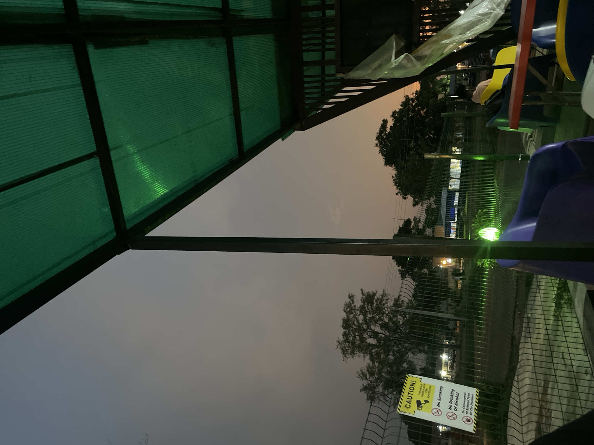 The image, rotated correctly for viewing, depicts an outdoor or semi-outdoor setting at twilight, likely in Ilorin, Nigeria. The perspective is looking upwards and outwards from beneath a large green corrugated plastic canopy, supported by dark metal beams, which occupies the upper left portion of the frame. A prominent dark metal beam runs vertically through the center of the image.
To the right, parts of brightly colored plastic seating, in shades of blue, purple, and yellow, are visible, suggesting an area for dining or recreation. A person is partially visible sitting on a yellow chair in the mid-ground. A transparent plastic sheet is draped over a structure in the upper right.
Beyond the immediate covered area, a wire mesh fence separates the foreground from the background, where silhouetted trees, distant artificial lights, and power lines are visible against an overcast, desaturated orange-grey sky.
Attached to the fence in the lower right is a yellow and white "CAUTION!" sign. The visible text on the sign reads: "CAUTION! THIS AREA IS UNDER 24HR CCTV SURVEILLANCE," followed by "NO SMOKING," "NO DRINKING OF ALCOHOL," and "NO CONSUMPTION OF EXTERNAL FOOD ON THE PREMISES." The scene is illuminated by dim ambient light and some artificial green lighting that reflects off the canopy and parts of the fence. No specific activity or event is distinctly taking place, conveying a sense of quiet observation.
