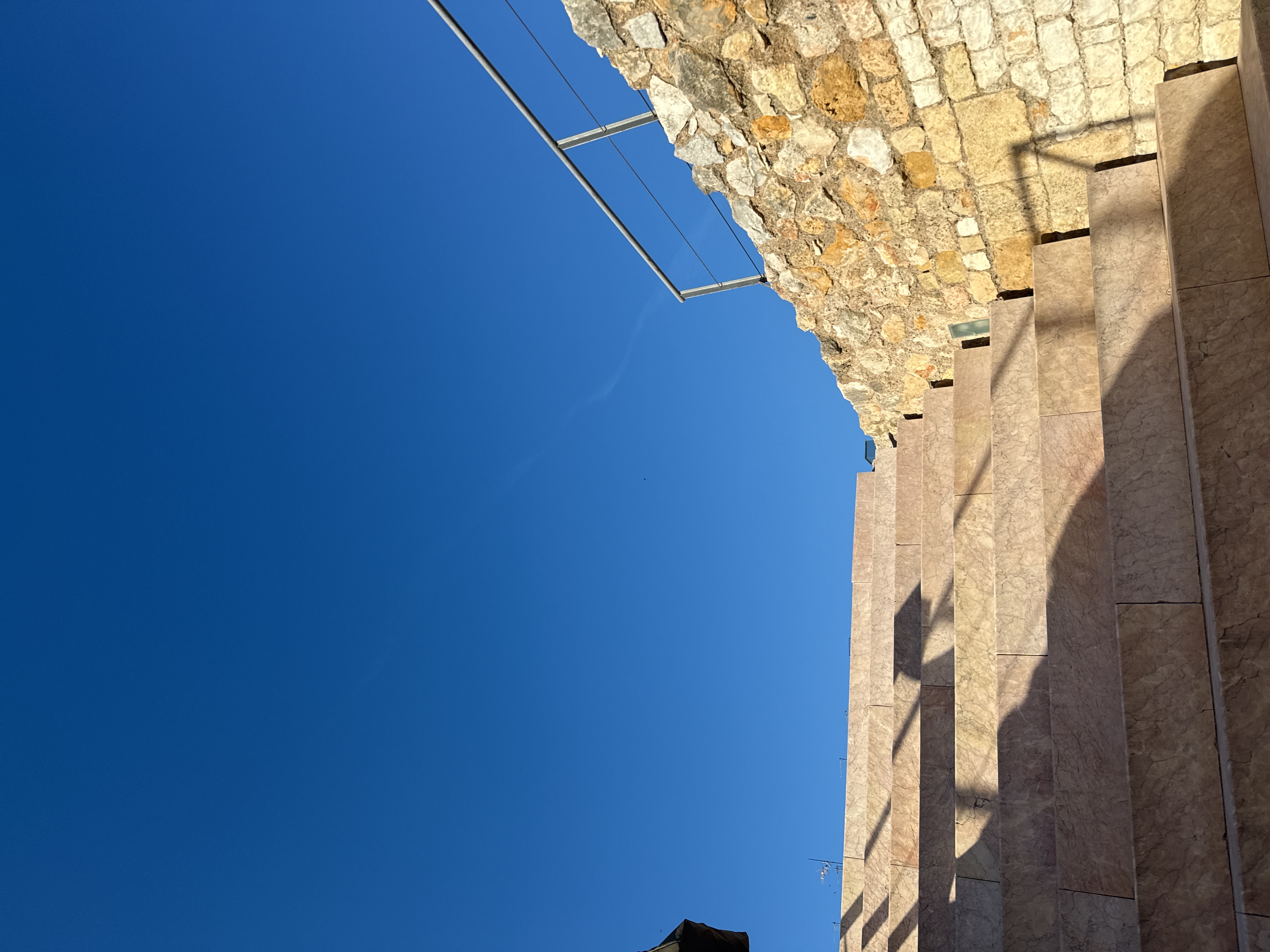 The image depicts a sunlit outdoor scene with a clear blue sky overhead. A set of stone steps rises diagonally across the lower half of the frame, casting shadows that add depth and texture. To the right of the steps, a rough stone wall is visible, topped with a simple metal railing. The architecture suggests a historical or public space.
Based on the architectural style and available information, it is likely that the location is in the city of Tarragona, Spain. However, definitive confirmation would require additional contextual information.