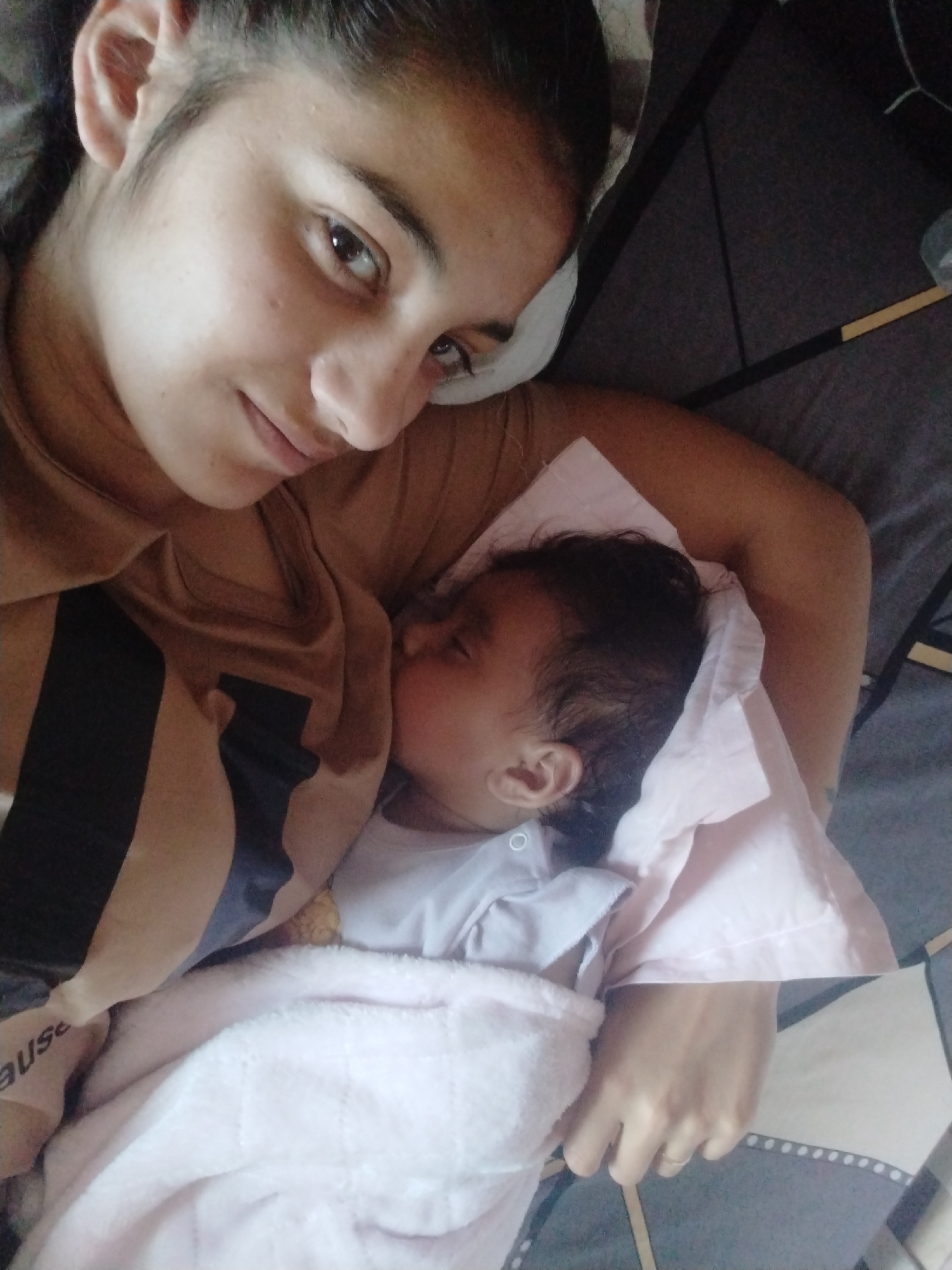A woman is lying on a bed, cradling a baby. The woman has dark hair pulled back from her face and is wearing a brown shirt with black stripes. The baby is wrapped in a pink blanket. The bed has a gray and white patterned cover. The location is Unknown, Unknown.