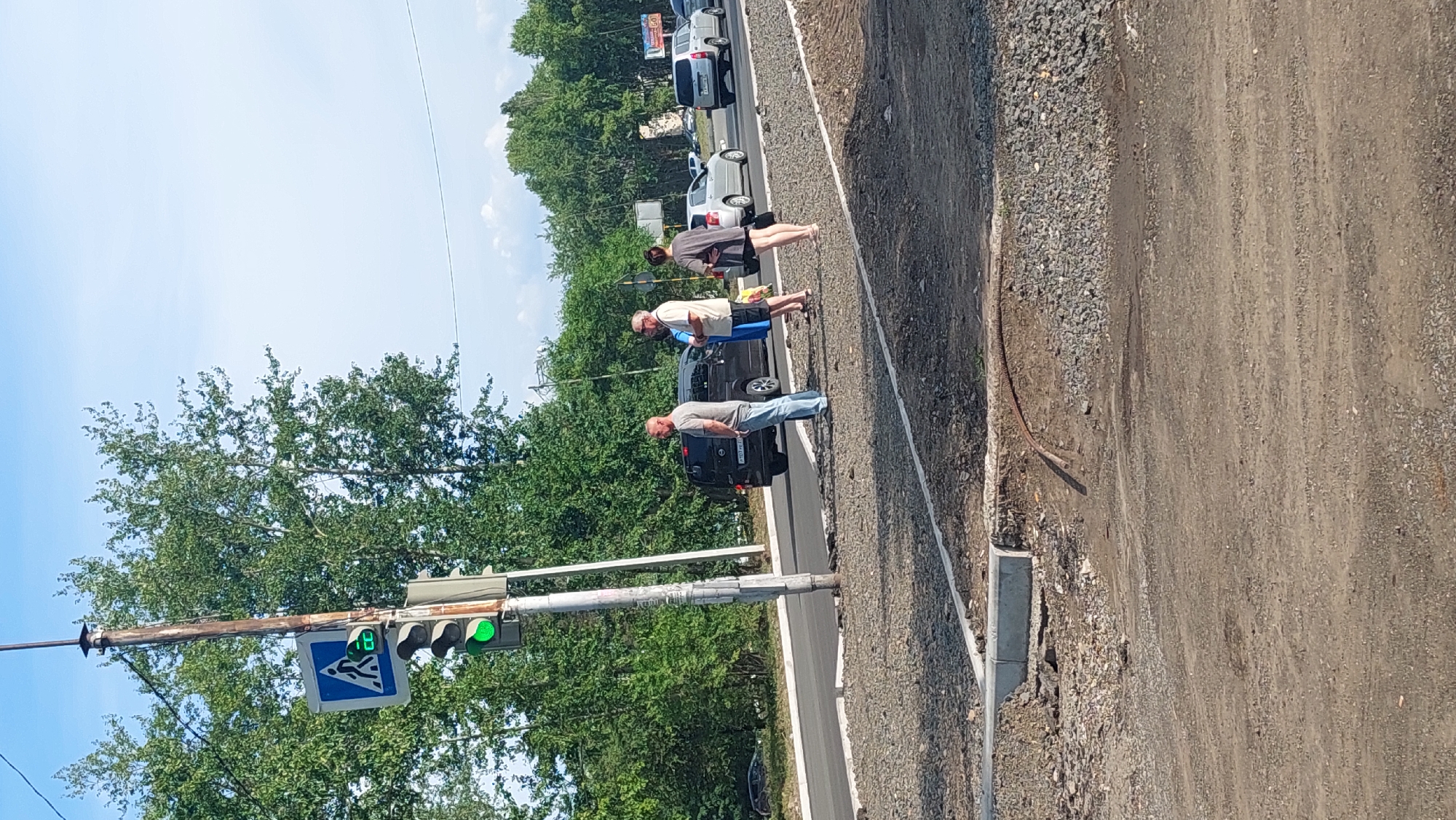 In Amursk, Russia, a traffic light on a utility pole displays a green light, indicating it is safe for pedestrians to cross. A blue pedestrian crossing sign is mounted above the traffic light. Three individuals are visible standing on the gravel shoulder next to the road. A dark-colored van is stopped on the road, and behind it, a silver car and a silver SUV are also present. The background consists of lush green trees, and the sky is a clear blue with some light clouds. The area appears to be undergoing construction, with loose gravel and some exposed dirt.
