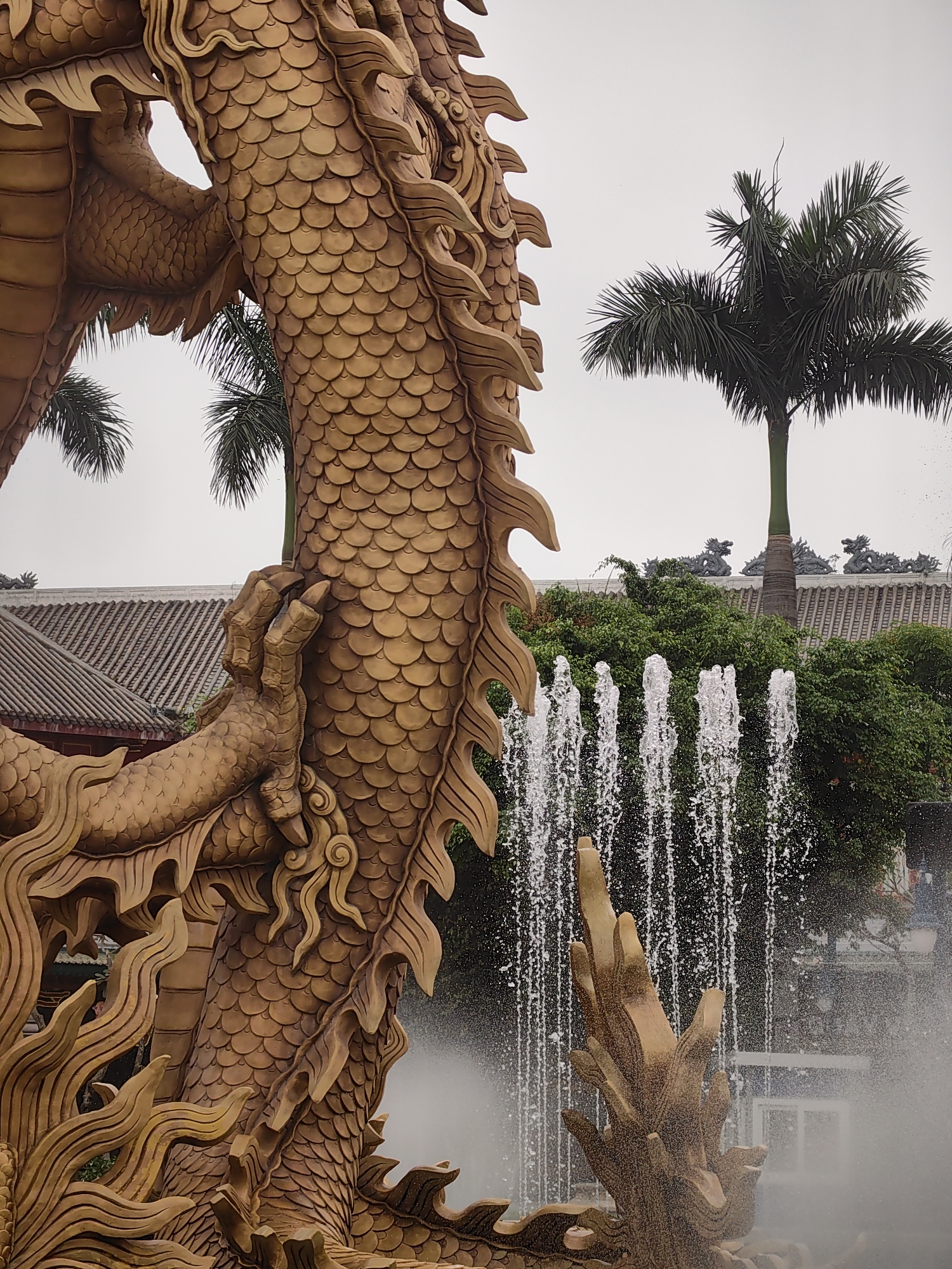 A large, ornate, golden dragon statue dominates the foreground, with detailed scales and flowing mane. The statue appears to be part of a fountain, as water jets are visible behind it. The background includes palm trees, green foliage, and a building with a traditional tiled roof. The presence of the dragon statue implies a cultural or religious significance, possibly located in a park or garden setting. The overcast sky contributes to the subdued lighting of the scene. The location is Bai Chay Ward, Vietnam.
