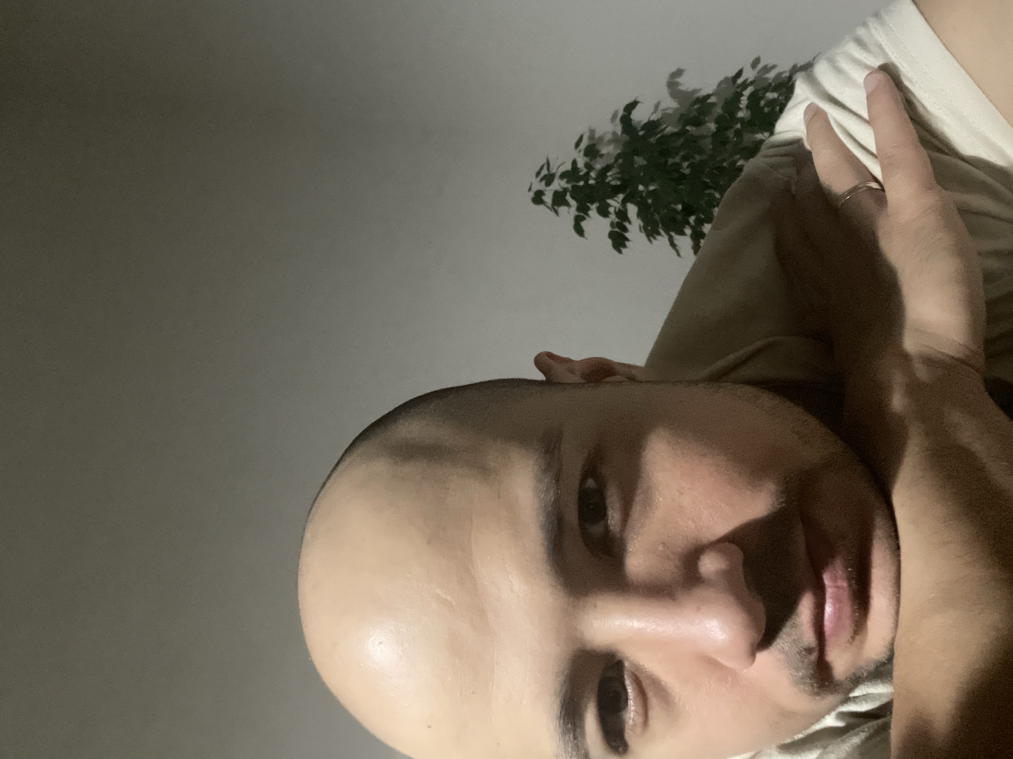 A close-up, low-angle shot of a bald man lying down. His face is partially illuminated, revealing features such as eyes, nose, and mouth. His hand is raised near his head. He is wearing a light-colored shirt. In the background, a green leafy plant is visible against a light gray wall. There is no evidence in the image to indicate a location in Izhevsk, Russia.