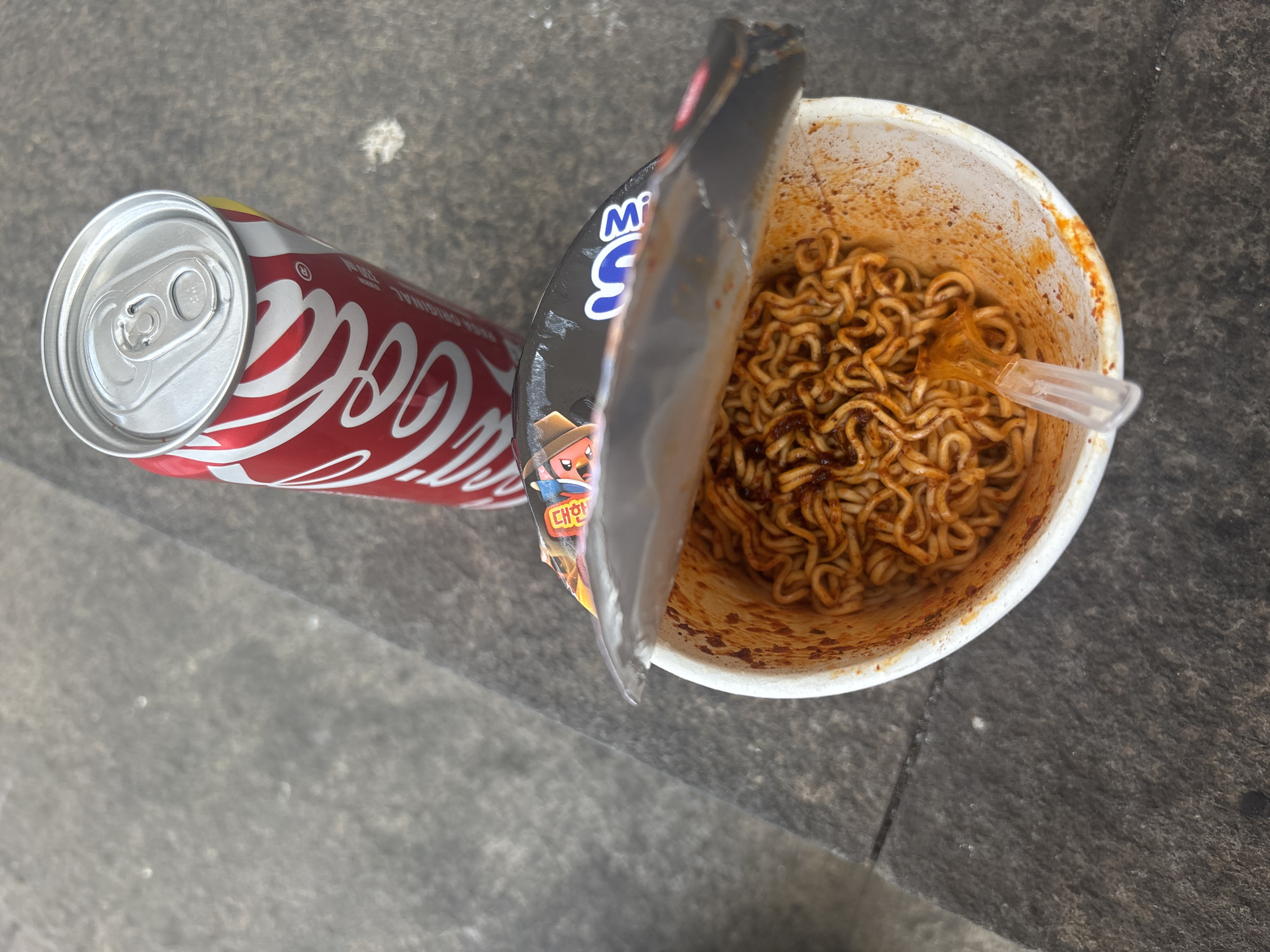 An overhead shot captures a Coca-Cola can and a cup of instant noodles on a grey, textured surface. The Coca-Cola can is red with white lettering and the iconic Coca-Cola logo. The cup of noodles is white and appears to be a spicy variety, with noodles coated in a reddish sauce. A clear plastic fork is visible inside the cup. The noodles are packed tightly and some sauce has splattered on the sides of the cup. The scene is lit by natural light, casting subtle shadows. The textured surface suggests an outdoor setting, possibly a park bench or a paved area.