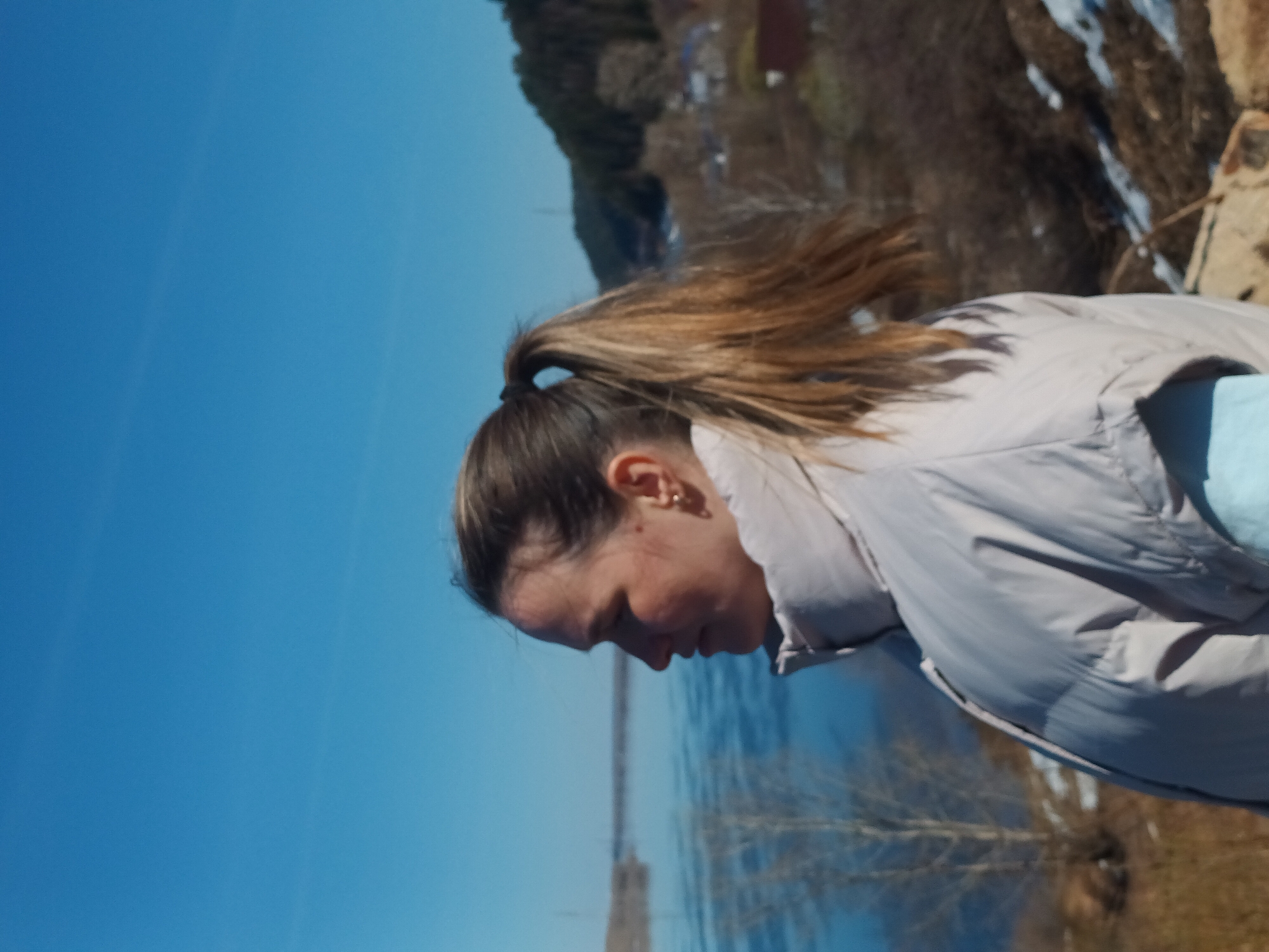 A medium shot depicts a woman with brown hair in a ponytail, wearing a gray puffer jacket and a light blue shirt, looking down. The background features a body of water, likely a lake or river, with trees and possibly buildings visible in the distance. The sky is bright blue. The location is possibly near the city of Ольховка, Russia, given the landscape and vegetation.