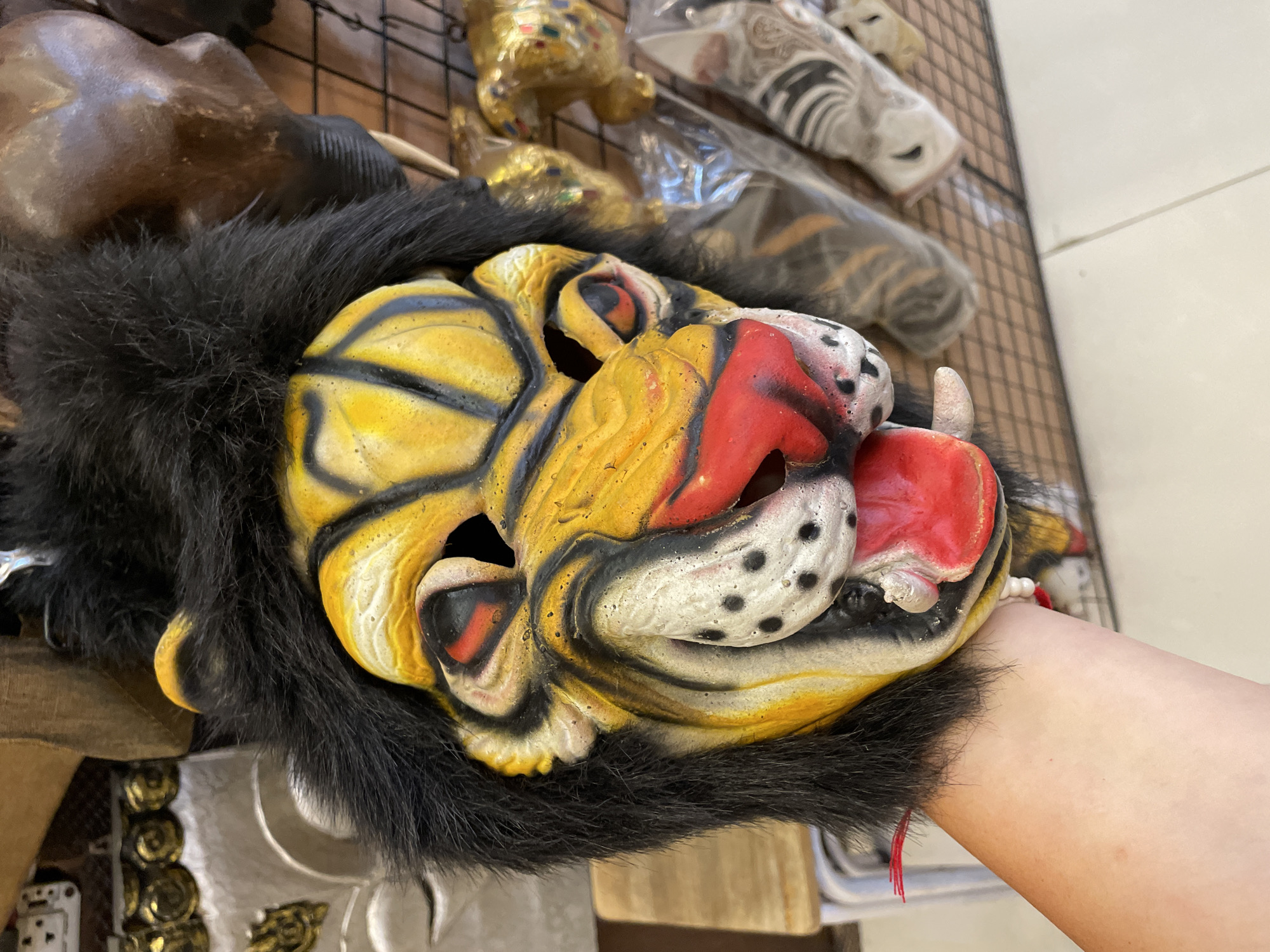 A person's arm and hand are holding a colorful, stylized lion mask in what appears to be a shop or market stall. The mask is predominantly yellow with black stripes, a red nose, and a wide, open mouth with a red tongue and white teeth. It has black fur around the edges. In the background, other masks and decorative items are visible, displayed on a black grid shelf. One of these items is a golden decorative piece with colorful accents, possibly also a mask or figurine. The floor appears to be tiled. The overall setting suggests a tourist-oriented retail environment. There is no specific visual information within the image to confirm the location as Pattaya City, Thailand, but the style of the masks and the general atmosphere are consistent with such a location.