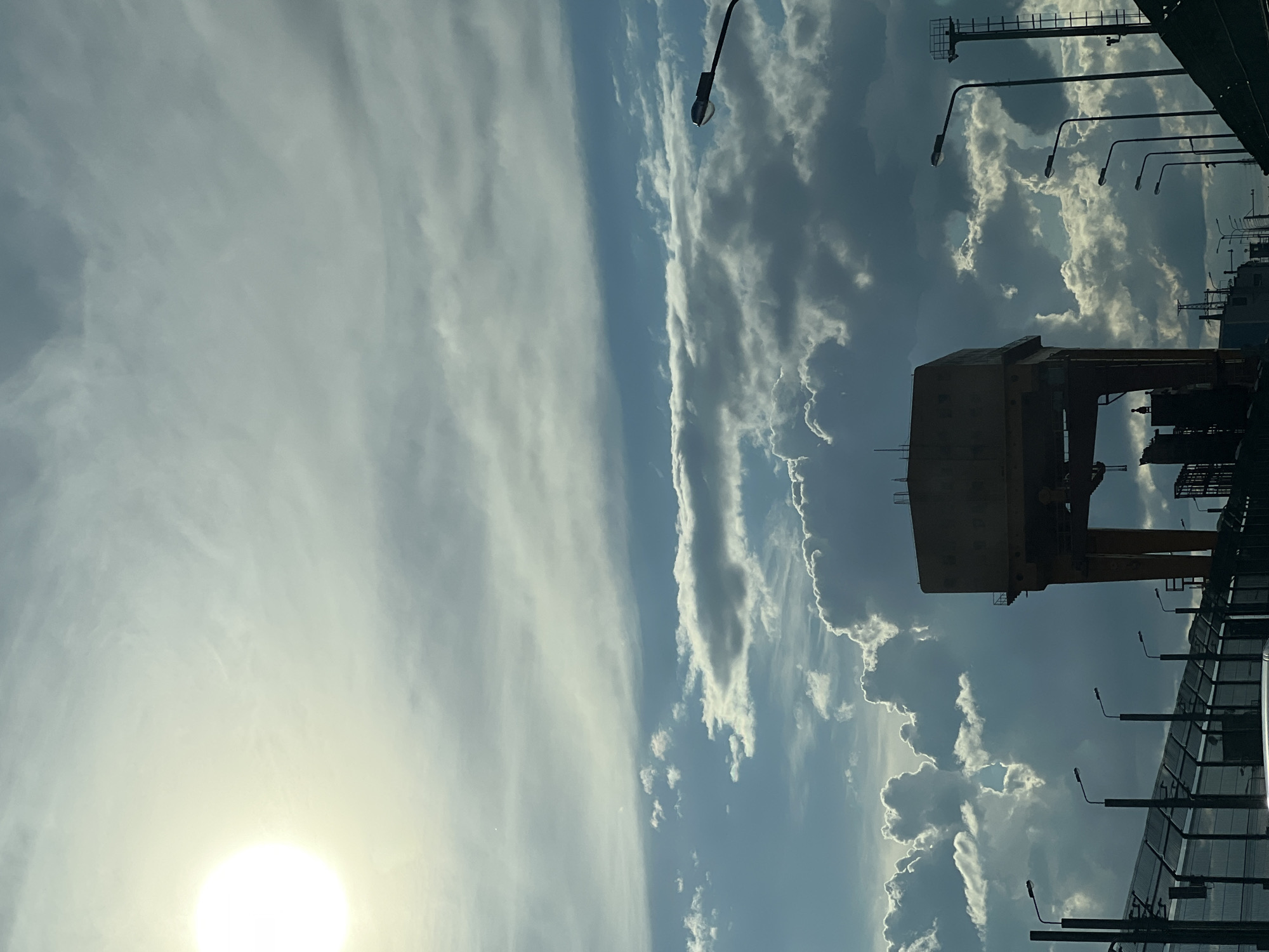 The image depicts a view in Chaikovsky, Russia, notable for its industrial landscape under a partly cloudy sky. In the foreground, a portion of a bridge or elevated structure is visible, lined with streetlights and a safety fence. To the right of the bridge, an industrial crane stands prominently against the sky. The sky occupies the majority of the image, with the sun shining brightly on the left side, and scattered clouds distributed throughout the blue sky.