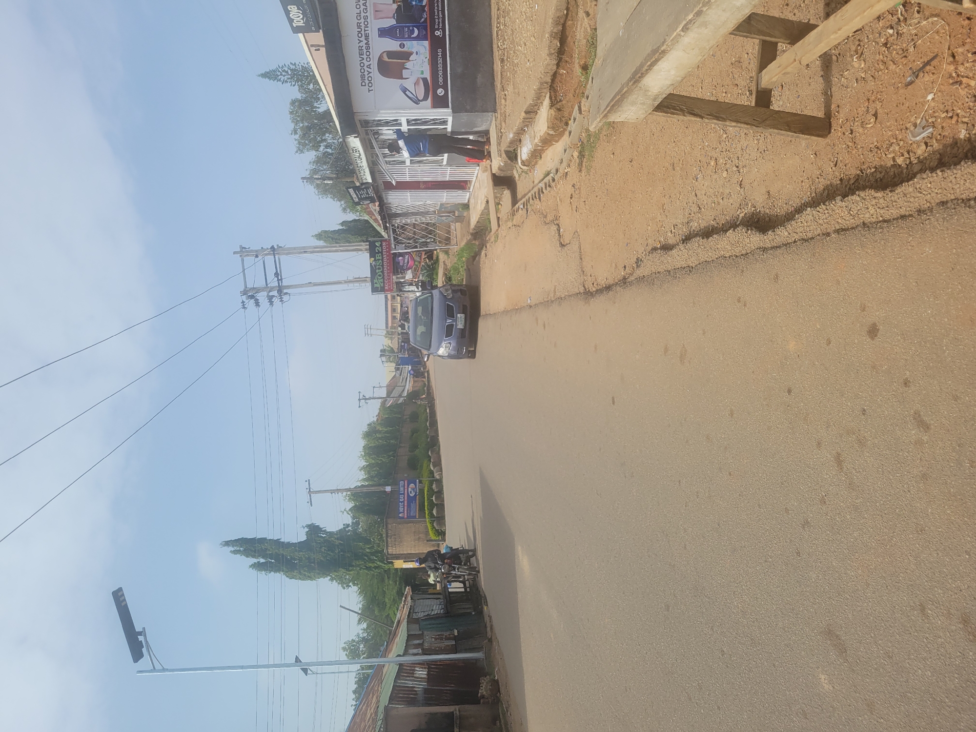The image captures a street scene in Jos, Nigeria. The asphalt road occupies a significant portion of the frame. A blue car is traveling down the road towards the viewer. A motorcycle with a rider is also present. On the right side of the road, a building houses several businesses, including "Tooya Cosmetics Gallery" and "House 24 Accommodation". A billboard for "ANEVIC GAS LIMITED" is visible further down the road on the left. Utility poles with power lines stretch across the sky. A modern street light stands tall on the left side of the street. A man is standing inside Tooya Cosmetics, and the scene suggests typical urban street activity.