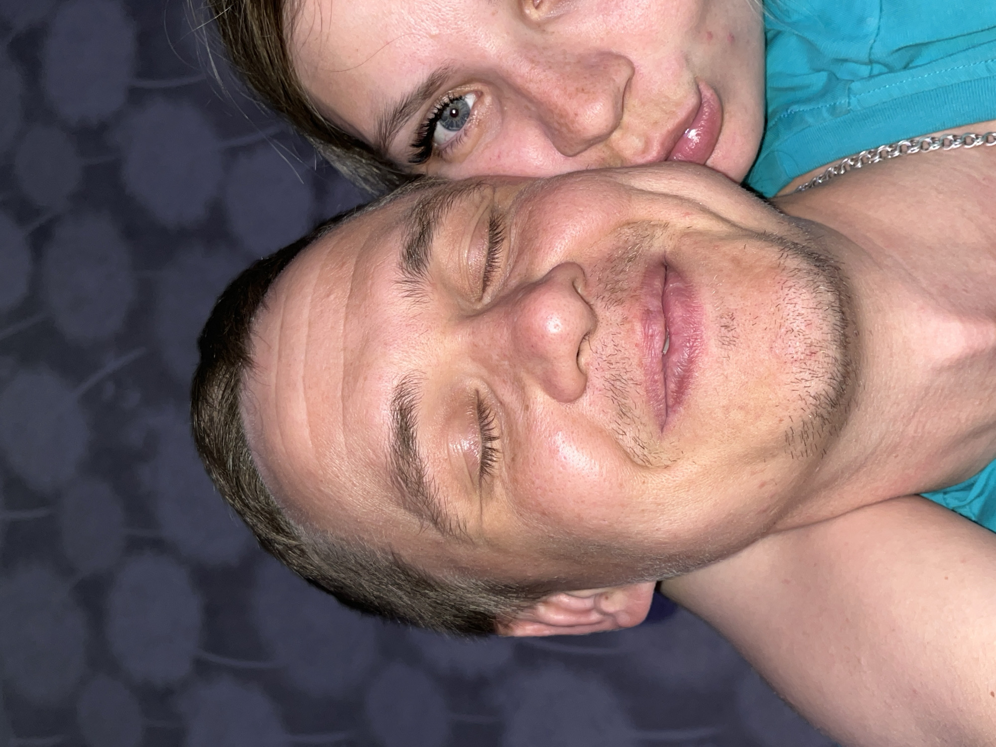 This intimate, close-up portrait features two individuals, a man and a woman, positioned in a tender embrace or resting posture. The image is oriented with the subjects horizontally in the frame, creating a sense of closeness.
The woman is visible in the upper part of the frame, resting her head on the man's shoulder or neck. She has light hair, fair skin with some freckles, and striking blue eyes that are open and looking towards the viewer. Her eyelashes appear long and defined, possibly with extensions. She wears a turquoise garment, likely a t-shirt, which is visible on the right side of the image.
The man occupies the lower and central part of the frame, lying with his eyes closed, suggesting he is sleeping or deeply resting. He has short, dark hair and light stubble on his face. A silver chain necklace is visible around his neck. His expression appears peaceful and relaxed.
The background is a dark, subtly patterned fabric, possibly a blanket or duvet cover, indicating an indoor setting, likely a bedroom or comfortable living space. The lighting is soft and ambient, suggesting it might be evening or night. No specific weather conditions are discernible. There is no visible text in the image.
The overall mood is one of comfort, intimacy, and affection, capturing a quiet moment between the two individuals. The location context provided is Chaikovsky, Russia.