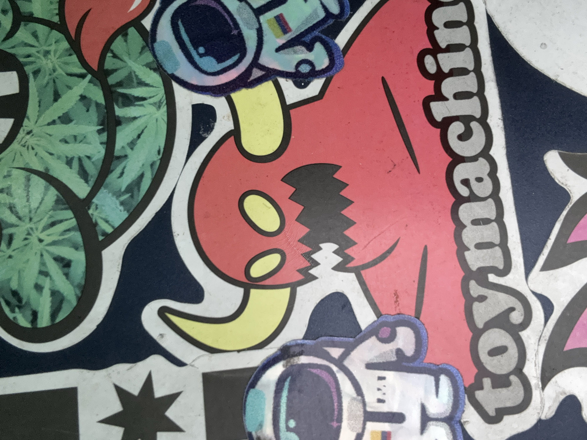 The image shows a close-up of stickers adhered to a dark blue surface. The stickers include a green cannabis leaf design, a red devil-like figure with yellow horns and eyes, two astronaut stickers, and a "Toymachine" sticker. The overall scene suggests a skateboard or other item commonly decorated with stickers. Without additional information, pinpointing the exact location in Cuautitlán Izcalli is impossible.