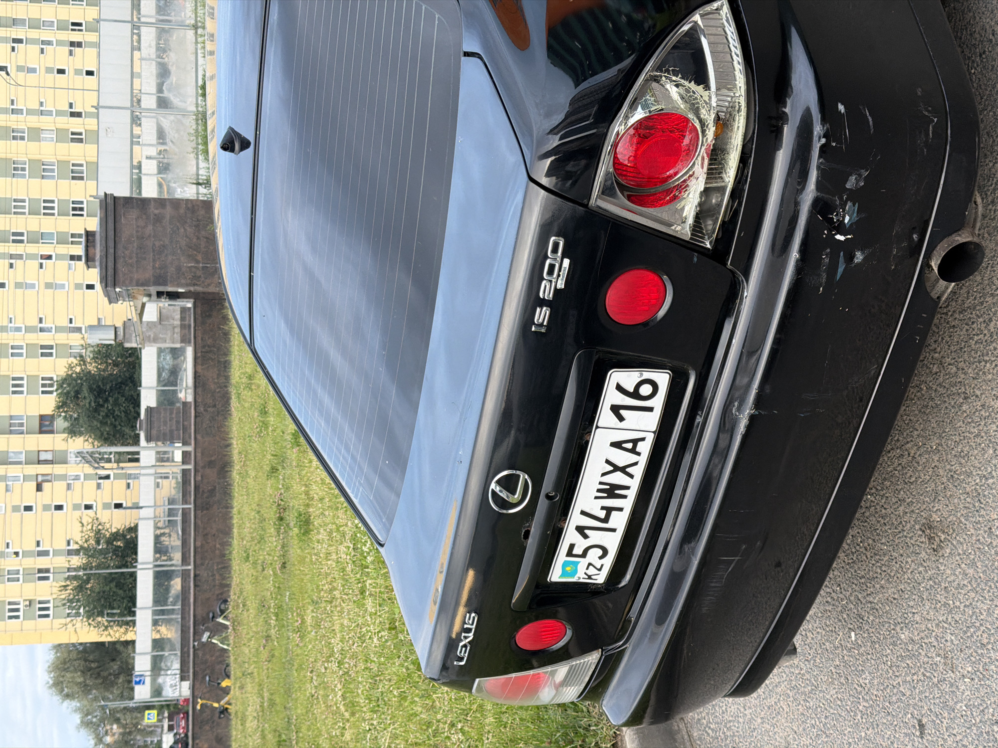 A black Lexus IS 200 is parked on asphalt. The car's rear is visible, showing a license plate with the characters "KZ 514WXA 16". The car has some visible damage on the rear bumper and a cracked taillight. In the background, there is a grassy area with two electric scooters, a low wall, and a tall yellow building with many windows. The sky is overcast.