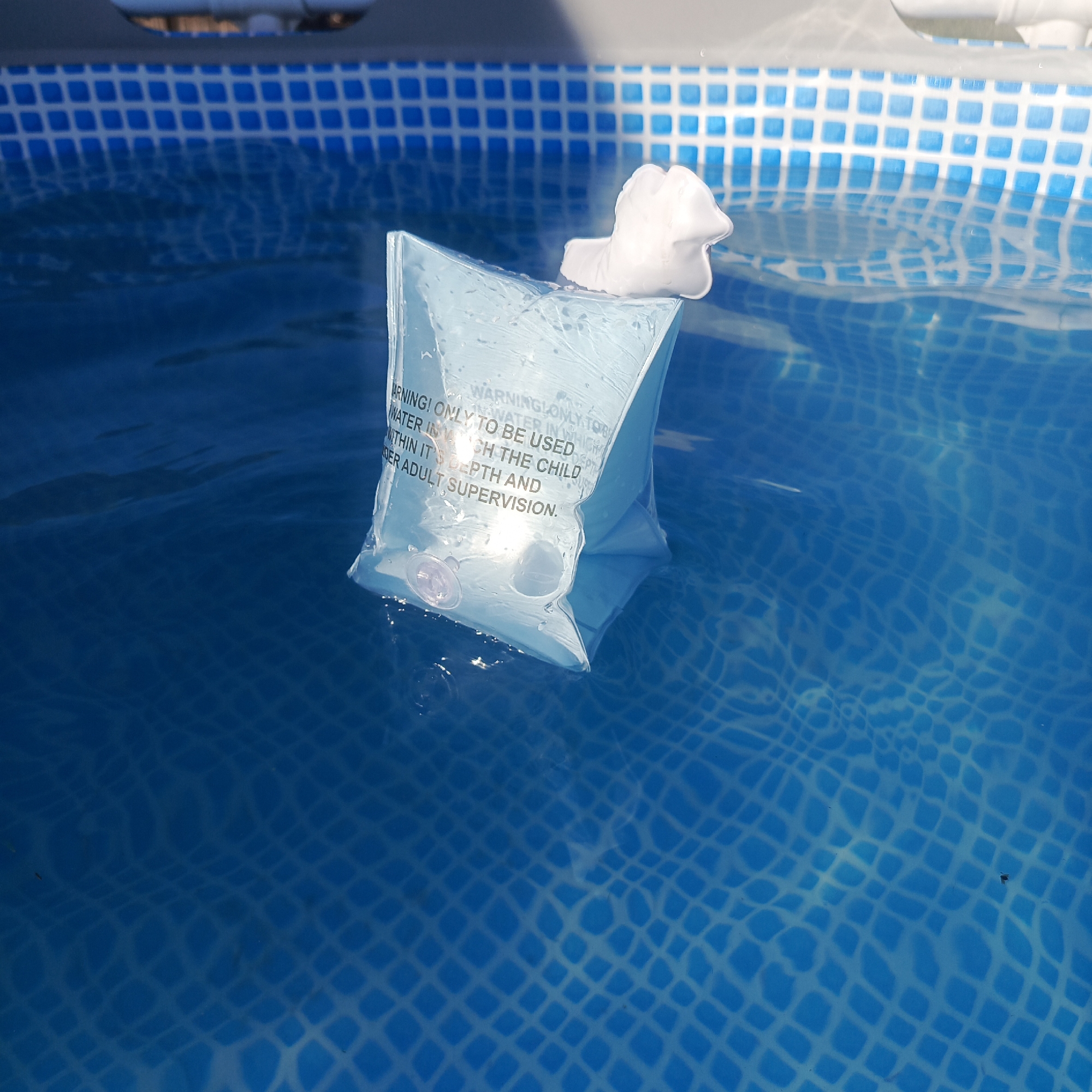 The image shows a blue and white tiled swimming pool containing water. An inflated, clear, plastic arm float, with a white foam pad attached to the top, floats in the water. The float has a printed warning message on its surface. The pool tiling can be seen behind the float. Ripples are visible on the water's surface.