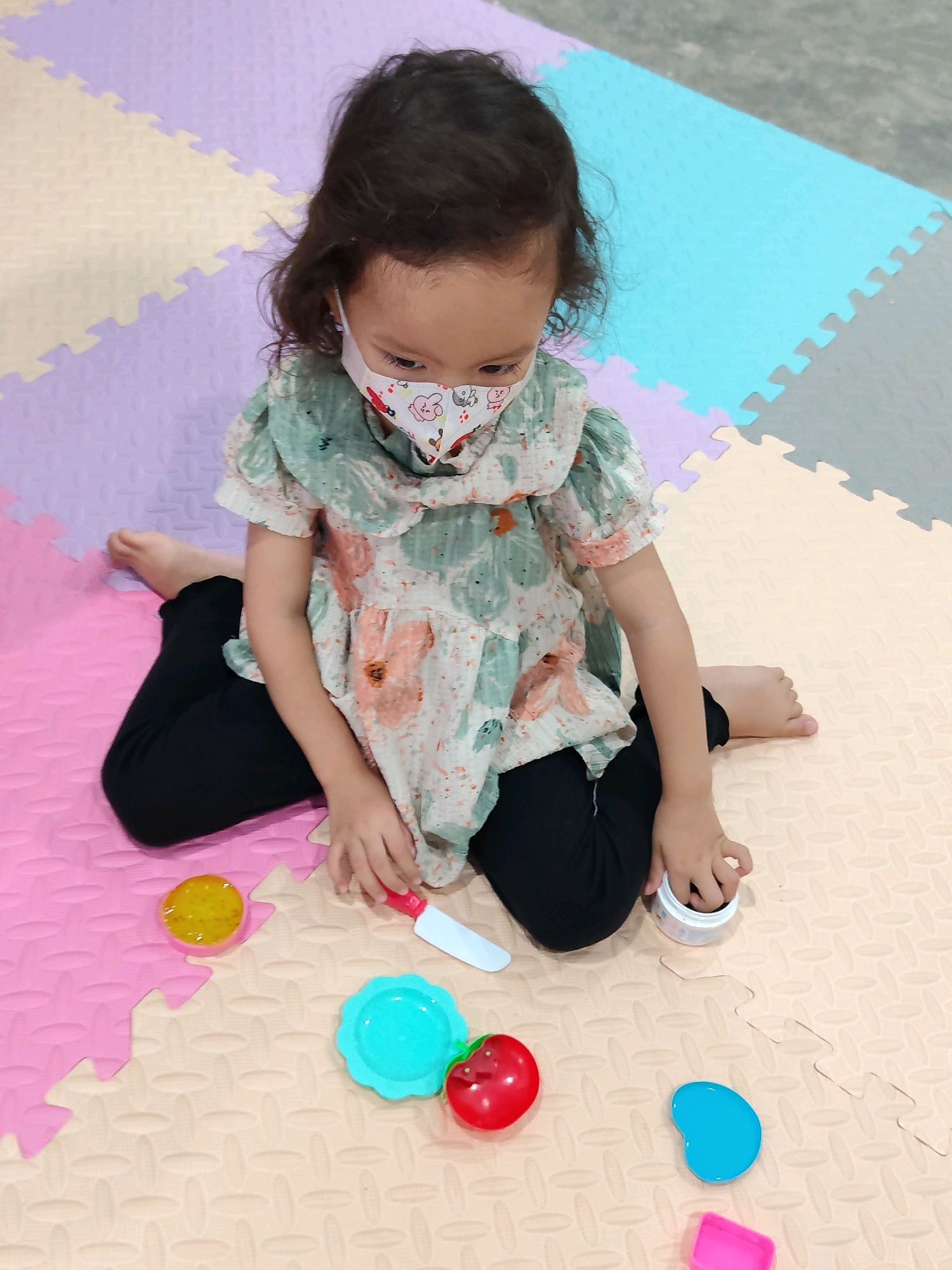 A young child wearing a floral print mask and a green and pink floral print blouse sits on a multi-colored foam mat, playing with toy food items. The child has dark hair and is wearing black pants. In front of the child are several toy food items including a yellow circular object, a blue plate, a red apple, and a blue bean-shaped object, as well as a pink rectangular object. A toy knife with a white blade and a red handle is positioned near the child's right hand. The child's right hand is also reaching into a small white container with a colorful lid. The mat is made of interlocking foam puzzle pieces in shades of pink, cream, light purple, and light blue, with some gray floor visible in the background. The scene appears to be an indoor play area, possibly in a residential setting.