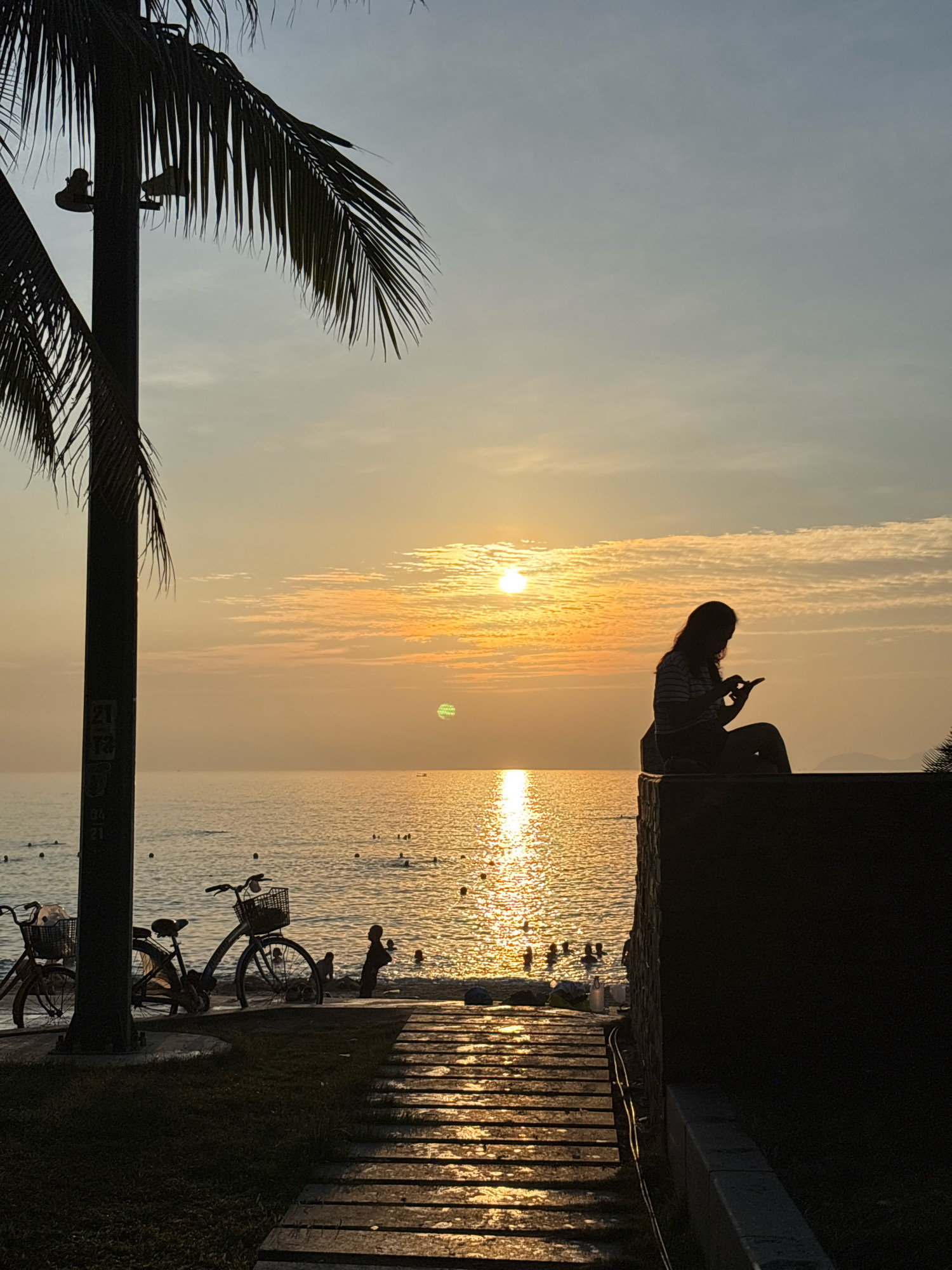 A silhouetted woman sits on a raised concrete structure, looking down at her phone, with the golden sunset reflecting on the ocean behind her. In the foreground, two bicycles with baskets are parked near a palm tree. The ocean is dotted with small figures of people swimming. A wooden walkway leads towards the water. The scene appears to be set at a beach in Nha Trang, Vietnam, characterized by its tropical coastline and evening twilight ambiance.