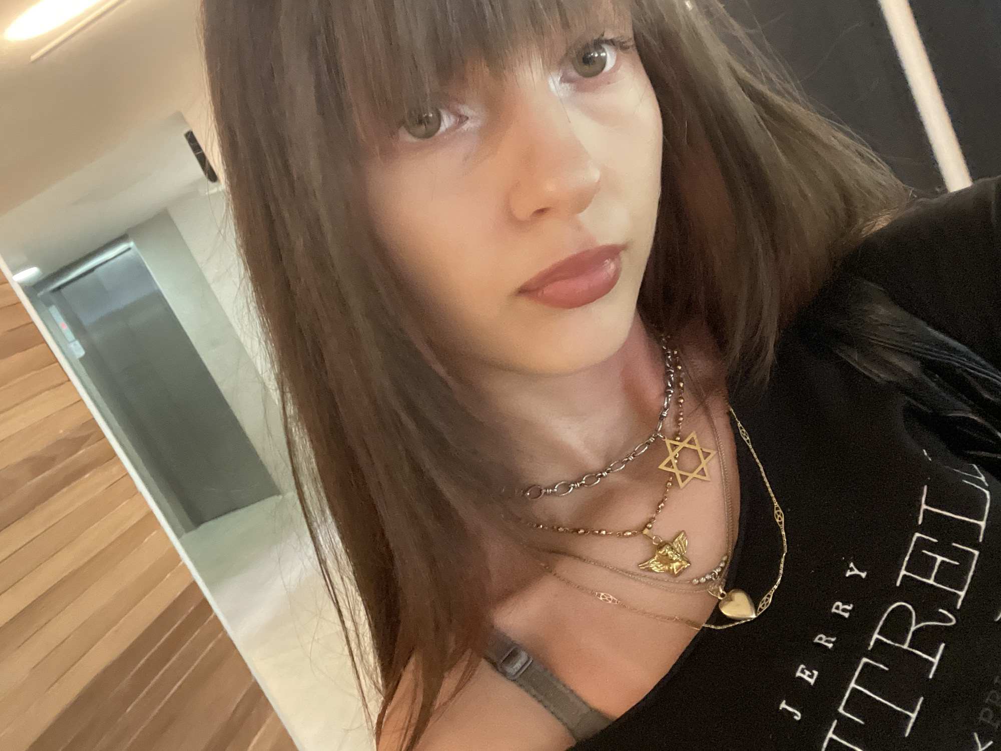 A medium, close-up selfie shows a young woman with long brown hair, bangs, and hazel eyes. She is wearing a black t-shirt with white text that reads "JERRY TRE..." partially obscured. Multiple necklaces adorn her neck: a silver chain, a gold Star of David, a gold cherub, and a delicate gold chain with a heart pendant. A grey bra strap is visible beneath her shirt.
The background shows part of an interior. To the left, there is a hallway with wood paneling on the wall and an elevator in the distance. To the right, a dark-colored wall is present. The precise location within Raanana, Israel, cannot be determined solely from the provided image.