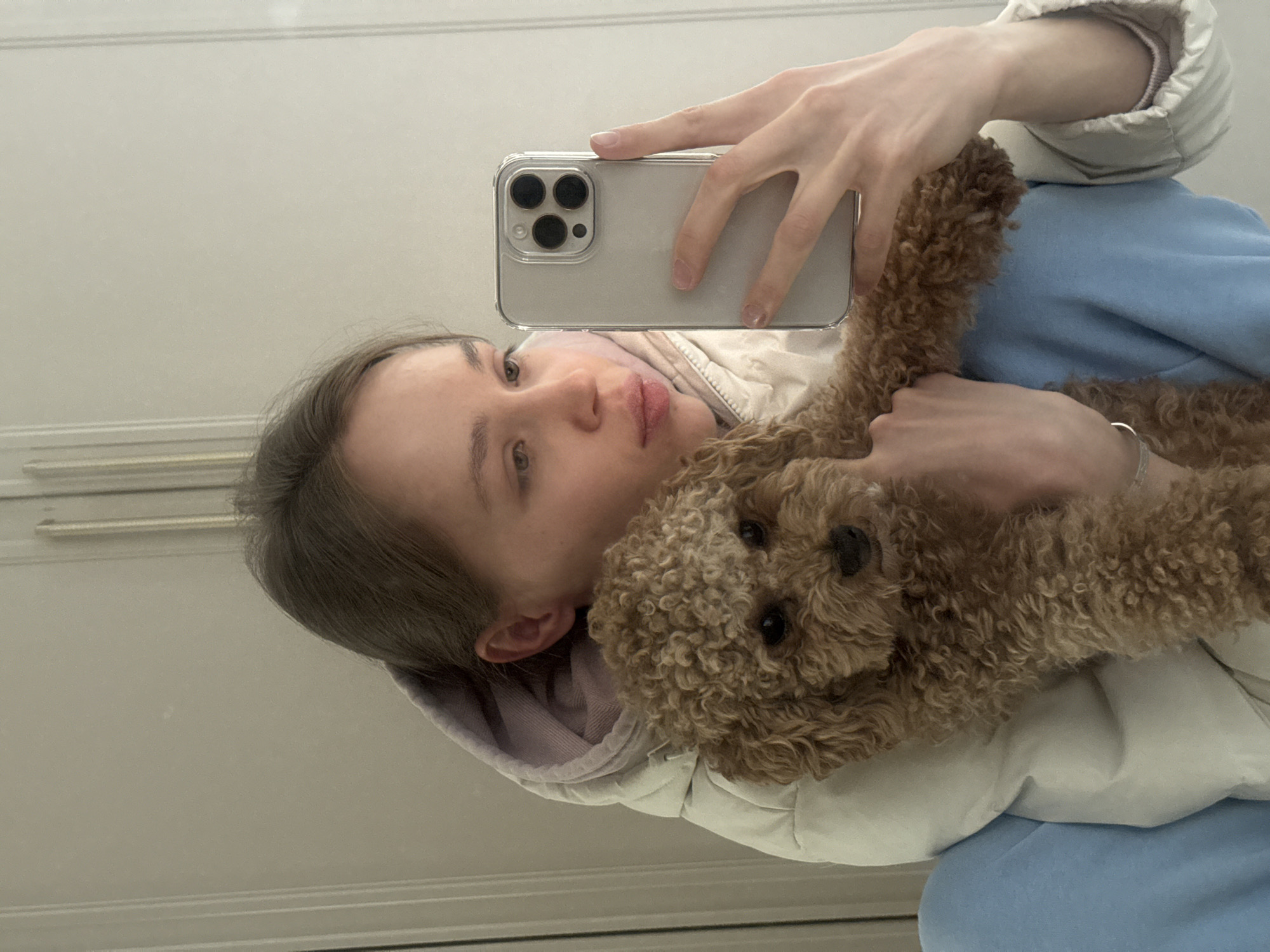A young person is taking a mirror selfie while holding a small, brown, curly-haired dog. The person, appearing young with light brown hair, has a neutral to slightly pensive expression. They are wearing a white puffer jacket over what appears to be a light purple hooded sweatshirt and light blue sweatpants. A silver bracelet is visible on their left wrist.
The person holds a silver or light-colored smartphone, likely an iPhone with a triple camera system and a clear case, in their right hand to capture the reflection. The small dog, resembling a poodle or cavapoo with its fluffy, brown fur and dark eyes, is held close to the person's chest and shoulder, looking towards the camera.
The background, visible in the mirror's reflection, consists of a plain, light-colored wall with subtle horizontal molding or trim. The setting appears to be an indoor environment, likely a home, with soft, even lighting. The overall image is rotated 90 degrees counter-clockwise. There is no visible text in the image.