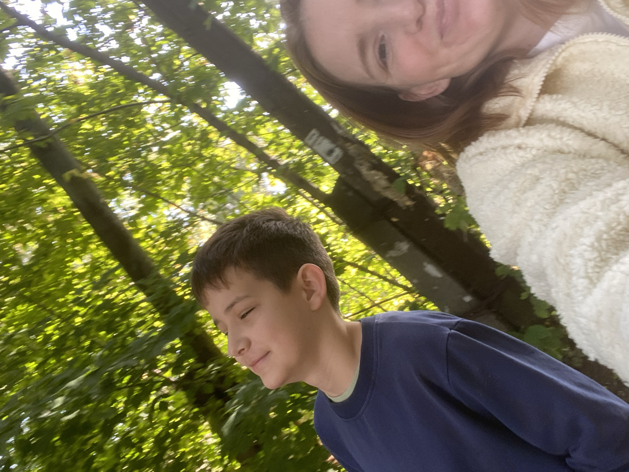 Two people are looking up into a canopy of trees, the sun dappling through the leaves. A boy, in the foreground left, wears a dark blue sweatshirt and has his eyes closed, tilting his head back. Beside him on the right, a woman's face is visible, smiling broadly, her head also tilted back. The background is filled with the blurred green of foliage and tree trunks. The setting appears to be a park or wooded area, possibly in Chaikovsky, Russia, given the information.