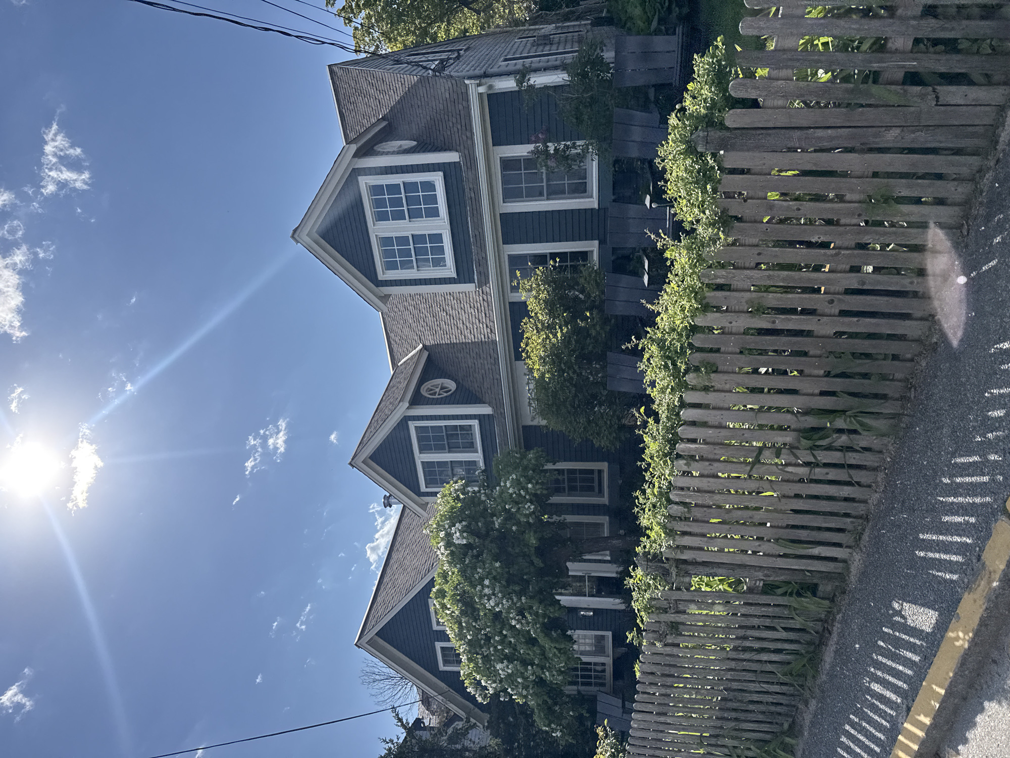 A three-story house is situated in Provincetown, Massachusetts, USA, viewed from a low angle. The house is painted a dark blue-gray with white trim and has multiple gabled roofs. The windows are multi-paned. A round window sits on the second story. The house is partially obscured by trees and foliage. A weathered wooden fence runs along the right side of the frame, with greenery growing along its top. A paved sidewalk is visible to the right of the fence. The sky is blue with some scattered white clouds. The sun is visible in the upper left corner, creating a lens flare. Power lines are visible in the upper left corner.