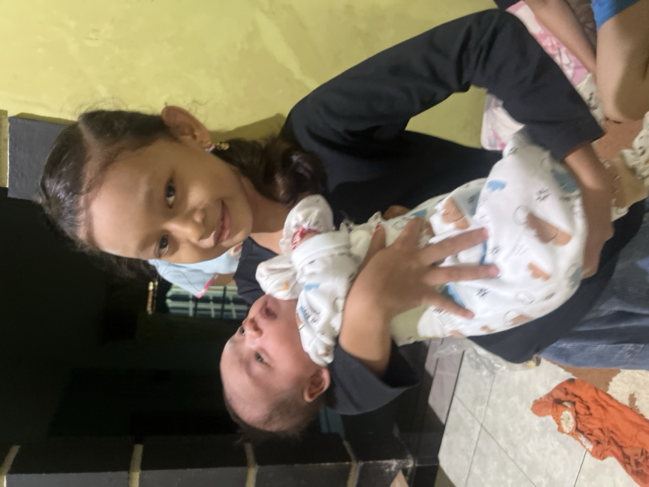 The image captures an indoor scene in Gabus, Indonesia, featuring two primary individuals. A female child, appearing to be approximately 8-12 years old, with dark hair partially pulled back, is positioned centrally. She wears a black long-sleeved shirt and blue lower garment, and small earrings are visible. She looks directly at the camera with a subtle smile. She is holding an infant, estimated to be under six months old, dressed in a white sleeper adorned with light blue and brown patterns. The infant, also with dark hair, is positioned with its head to the left, looking upwards and away from the camera. The girl's hands are seen supporting the infant. In the background, a light yellow-colored wall is visible on the right side of the frame, contrasting with a dark-colored structural element, possibly a pillar or wall corner, on the left. Partially visible behind the girl's left shoulder is another individual wearing a light-colored garment. The floor, visible in the lower portion of the image, is tiled, and an orange textile item lies on it in the bottom left. A pink and white patterned fabric, possibly a cushion or blanket, is partially visible in the lower right corner, adjacent to a leg clad in blue fabric.