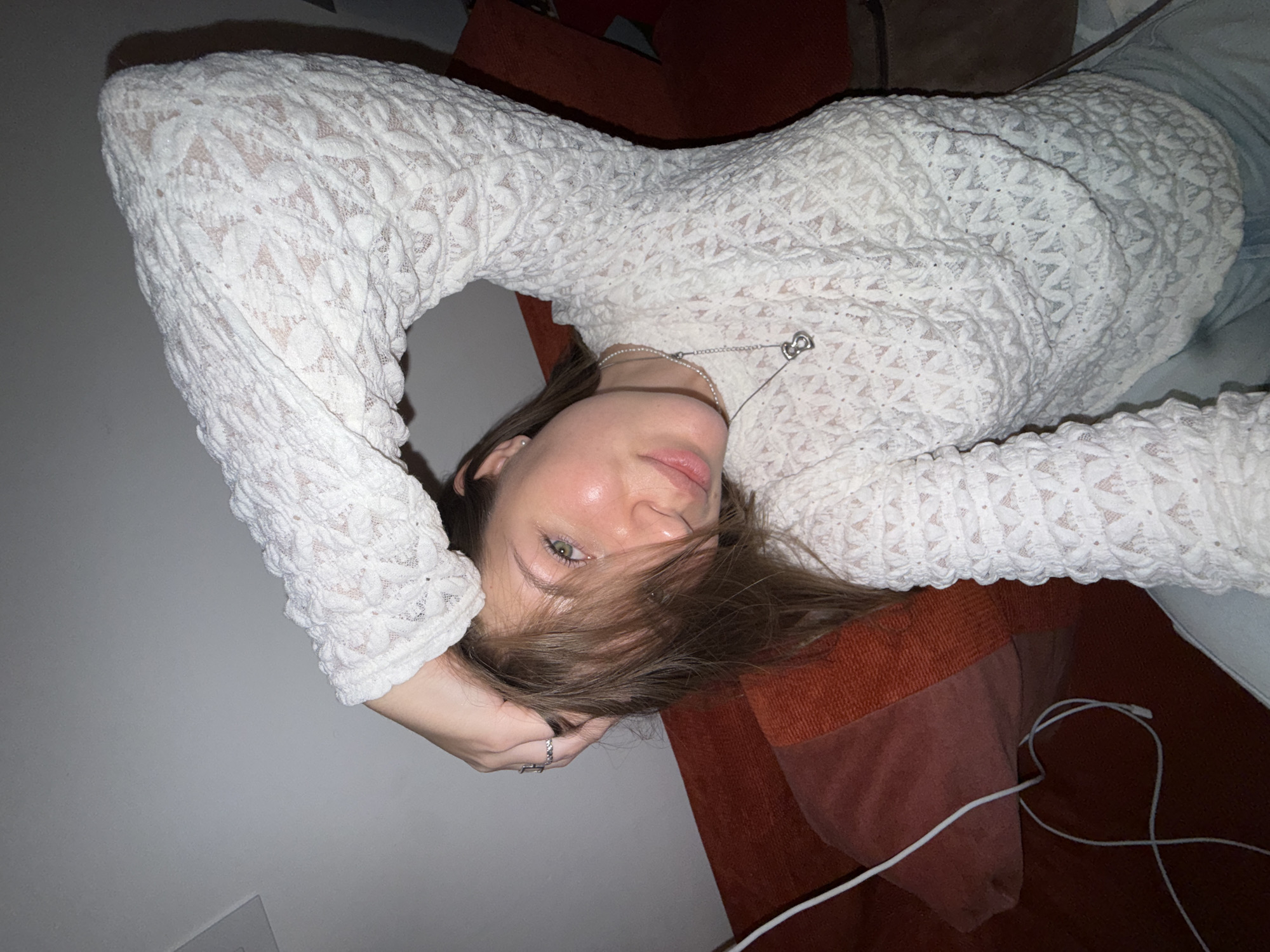 A young woman lies on an orange sofa with a white wall in the background. She is wearing a white, long-sleeved lace top and a silver necklace. Her hair is brown and partially covers her face. One of her hands, adorned with a ring, rests behind her head. A white charging cable is visible near the bottom of the image. The location is identified as Vozdovac Urban Municipality, Serbia, based on image analysis and potential correlation with geographical data, although specific indoor locations are not identifiable from visual data alone.