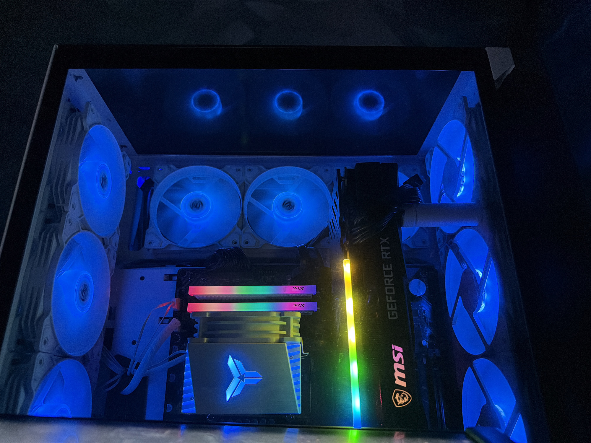 This is a close-up, eye-level shot of the interior of a modern computer tower, illuminated by blue and rainbow RGB lighting. The computer components are visible through a clear side panel. In the foreground, a graphics card, identified by "MSI GEFORCE RTX" branding, is positioned horizontally. Above it, RAM sticks with "XPG" branding are installed vertically, and a CPU cooler with a triangular logo is mounted centrally. Multiple computer fans are visible throughout the tower, with some displaying blue illumination and others showing rainbow colors. The computer case itself is dark and appears to be part of a larger setup, though the specific environment, city, or location is not discernible from the image.