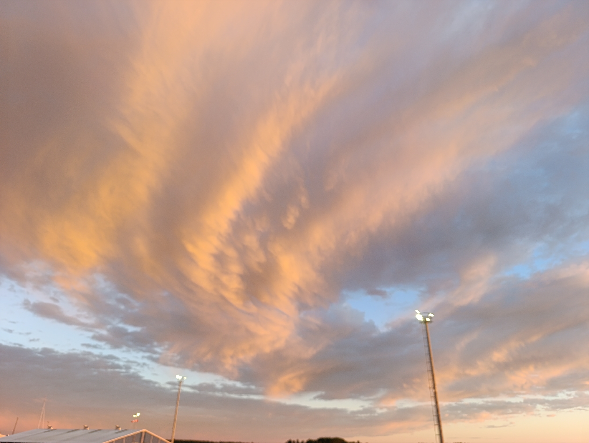 The image captures a sunset over the city of Jakobstad, Finland. The sky is filled with dramatic clouds displaying hues of orange, pink, and gray, indicating the last moments of daylight. In the foreground, the top of a building with a white roof is visible, along with a tall lamppost with illuminated lights. The overall impression is one of a serene evening sky over an urban landscape.