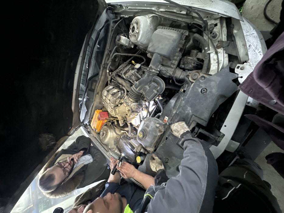 In Zhabkino, Russia, an elevated view captures an indoor scene within a garage or workshop. The primary subject is the open engine bay of a light-colored vehicle, revealing a somewhat soiled internal combustion engine and associated components. A red and yellow portable power or jump starter unit is visible on the passenger side of the engine bay.
Four adult males are engaged with the vehicle. In the center-left, a male with short dark hair and a beard, wearing a dark shirt with neon green trim, holds a black flashlight, directing its beam into the engine's deeper sections. Immediately to his right, another male, partially obscured, is observing. Above them, a male with a short dark beard, glasses, and wearing a dark, fur-hooded jacket over a light scarf, holds a lit cigarette between his fingers near his mouth as he looks down into the engine bay. In the lower-left foreground, a fourth male, identifiable by his gray work garment and beige work gloves, has both hands positioned on a black plastic shroud covering the front of the engine bay. The combined actions indicate a collaborative inspection or diagnostic process of the vehicle's engine. The environment is illuminated by artificial light sources.