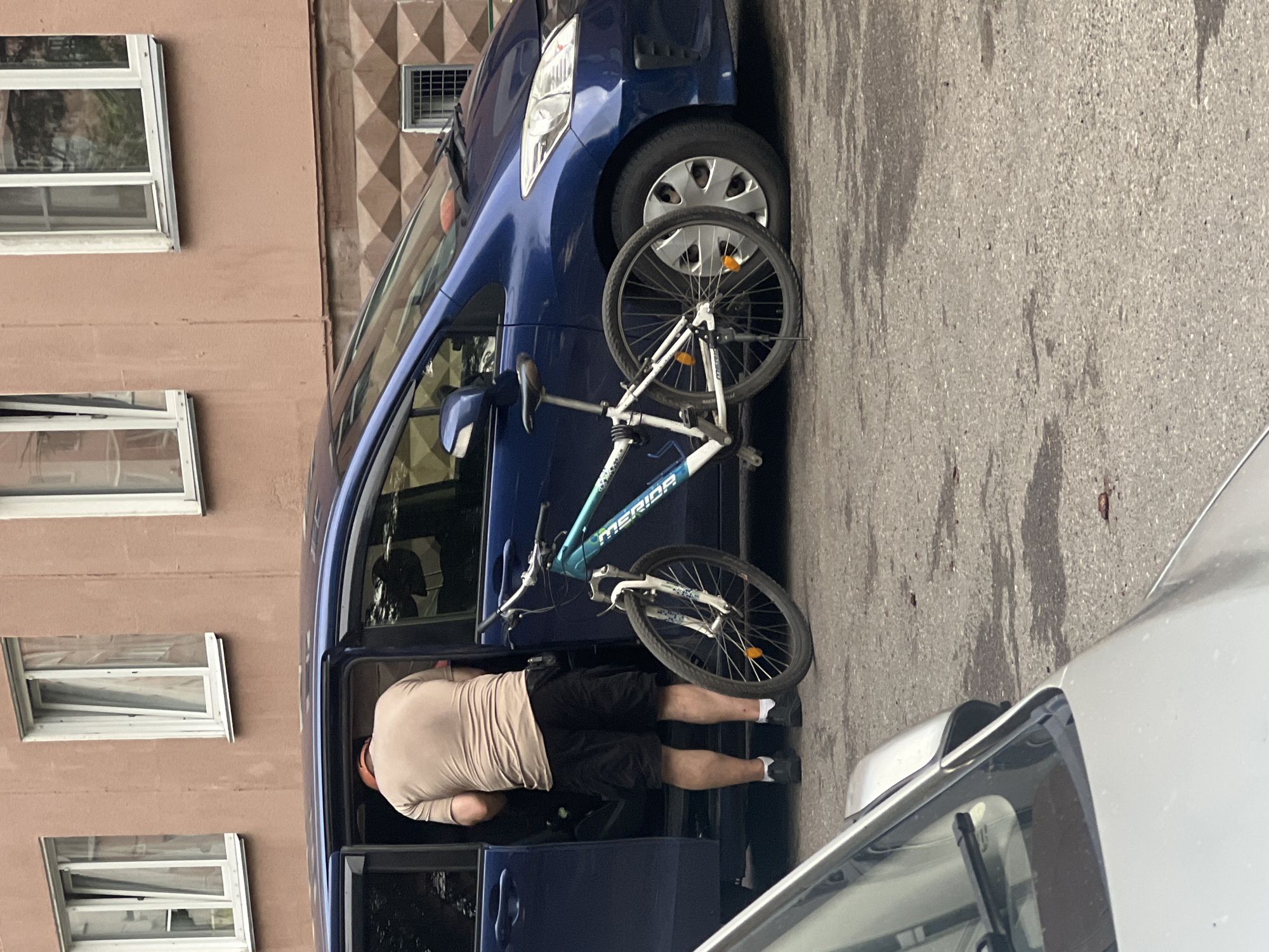 The image shows a man in black shorts and a light brown t-shirt bent over inside a blue car with the driver's side door open. A white and teal bicycle is propped against the car, with its handlebars and front wheel resting on the ground. The bicycle has "MERIDA" written on its frame. The man appears to be either loading or unloading the bicycle from the car.
The scene takes place on an asphalt surface, possibly a street or parking lot, next to a building with pinkish-brown walls and several windows. The building has a distinctive patterned facade above the windows. The time of day appears to be daytime, with overcast lighting. There are reflections in the car windows, including what looks like another person inside the car. A portion of a silver car is visible in the foreground. No explicit emotions are visible, but the posture of the man suggests he is engaged in a task.
The location context provided is Zheleznogorsk, Russia.