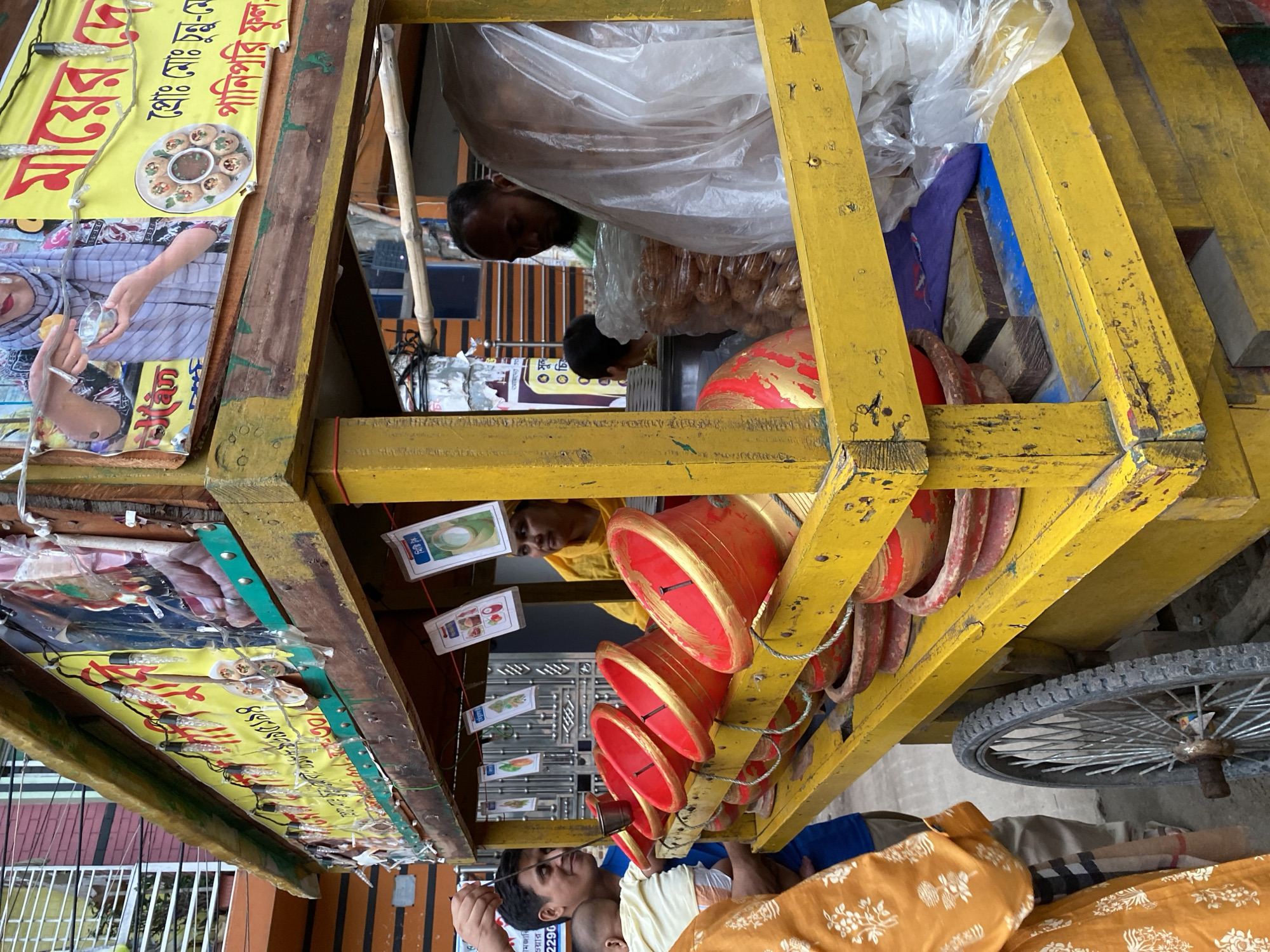 This image captures a vibrant street food scene in Dhaka, Bangladesh, centered around a brightly painted yellow wooden pushcart. The cart, equipped with a wheel visible on the right, functions as a mobile food stall.
Several people are visible in the frame, primarily men involved in the food preparation and selling. One man, partially obscured by a large clear plastic bag filled with what appears to be fried snacks (likely "fuchka" or "pani puri"), is leaning into the cart. Another man is seen from the chest up, looking outward, possibly attending to customers or preparing food. Underneath a row of red containers, another person's head is visible, and at the bottom right, a person in an orange patterned garment is present.
The cart is adorned with various objects related to its purpose. A prominent yellow poster on the left side displays a plate of round, fried snacks with a dark sauce, characteristic of "Pani Puri" or "Fuchka." Bengali text is clearly visible on this poster, alongside an image of a woman's hands holding a small cup and a piece of the snack. A string of small light bulbs is draped along the edge of the cart, indicating it might also operate in the evenings.
A row of red, bowl-shaped containers with gold rims is stacked and arranged within the cart, likely used for serving or holding ingredients. Small white cards featuring images, such as a green mango, and Bengali text hang from wires, possibly indicating menu items or ingredients. Inside the cart, various wooden planks, some painted blue, are visible, contributing to the cart's structure.
The scene depicts a bustling daytime environment. The natural lighting suggests it's a clear day. The Bengali script on the posters and the specific type of street food are strong indicators of the location being Dhaka, Bangladesh. The overall atmosphere conveys the routine activity of a street food vendor, with an emphasis on local flavors and a casual, outdoor setting. A partial phone number, "2290," is also visible on one of the signs.
