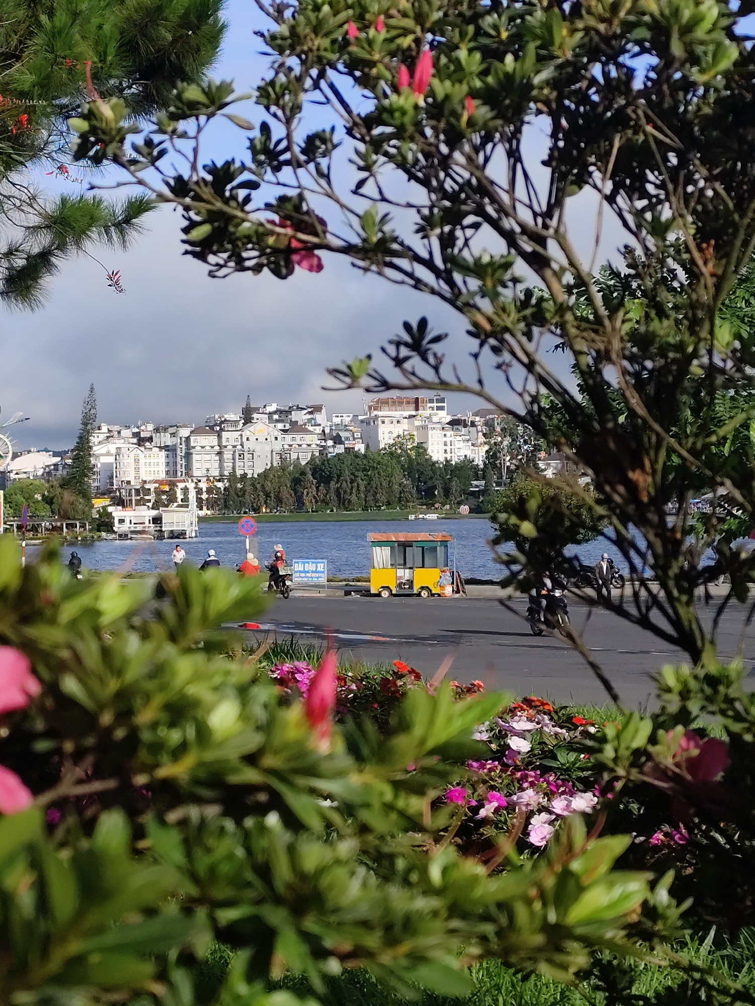 The image depicts Xuan Huong Lake in Da Lat, Vietnam, viewed from a foreground of lush green foliage and pink flowers. In the middle ground, a body of water is visible, with several people riding motorcycles along the paved lakeside road. A yellow food cart is parked on the side of the road, and a sign with Vietnamese text is posted nearby. In the background, a cityscape of buildings and trees lines the far side of the lake, under a bright, partly cloudy sky. The overall scene is a picturesque snapshot of a vibrant public space in Da Lat.
