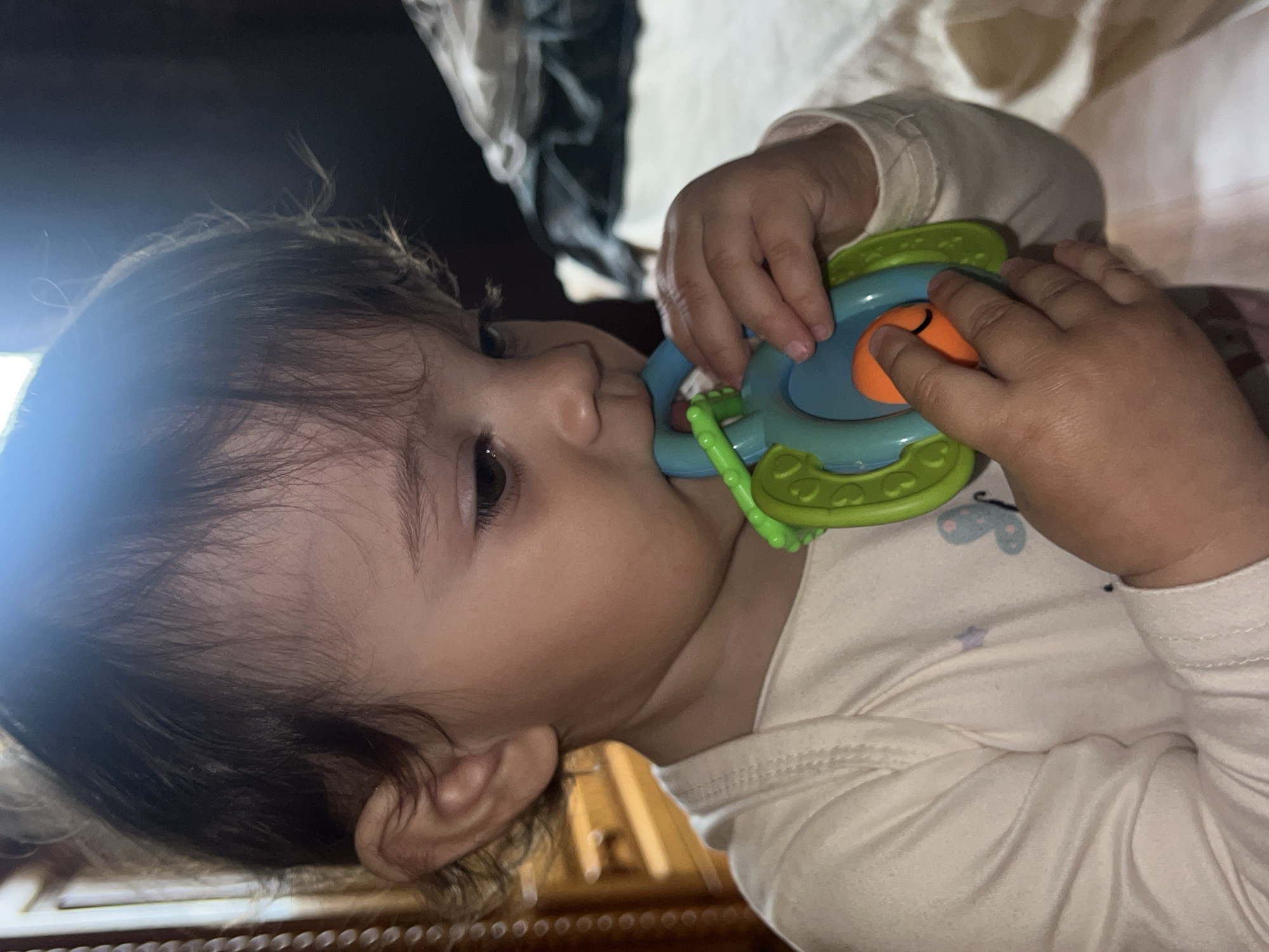 A baby is lying on a surface, possibly a bed or changing table, while holding a colorful toy. The baby is wearing a light-colored long-sleeved garment. The toy is round and features green, blue, and orange parts. The background is slightly blurred, showing light-colored fabric and possibly a dark object in the distance. The image is rotated counter-clockwise. There is no indication of location in the city of Elena, Bulgaria.